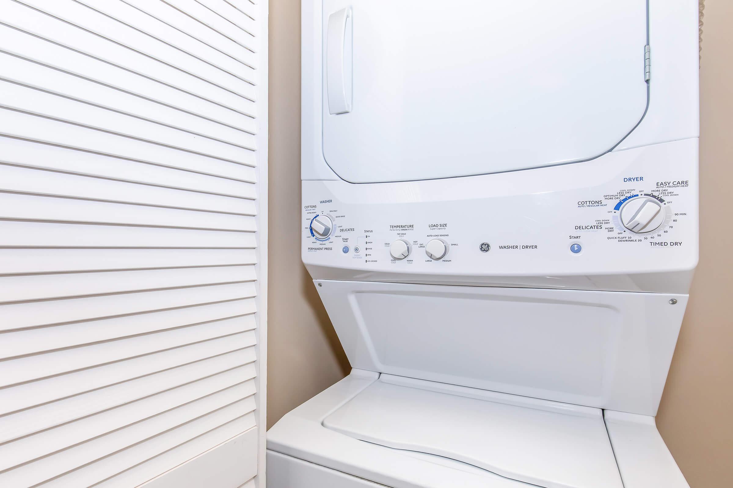 A compact washer and dryer unit stacked in a laundry room, featuring various control dials and settings. The unit is situated beside a closed louvered door, with a neutral-colored wall in the background.