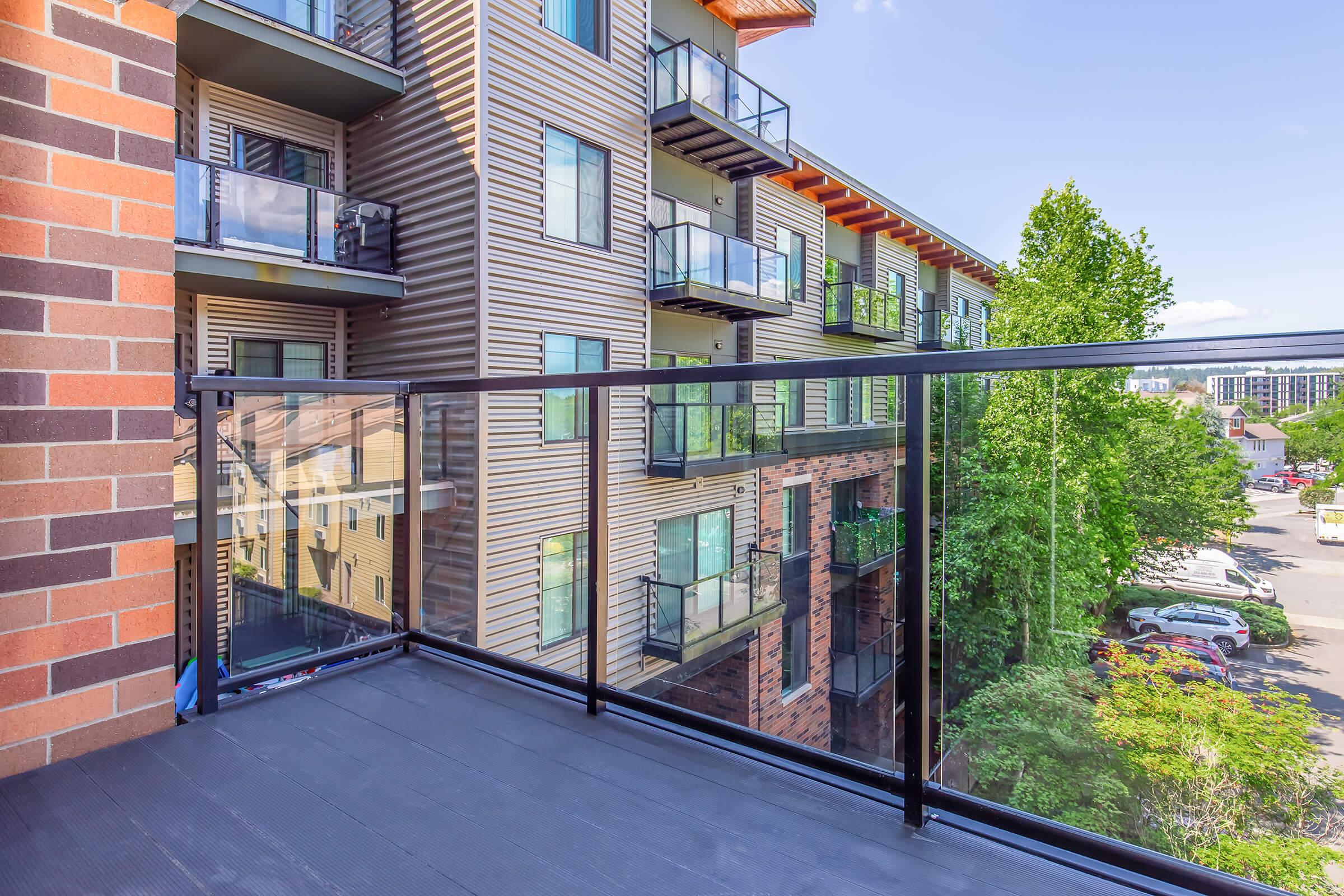 View from a balcony overlooking a multi-story apartment building with modern architecture. The balcony features glass railings, and there are trees and cars visible in the background, creating a mix of urban and natural scenery.