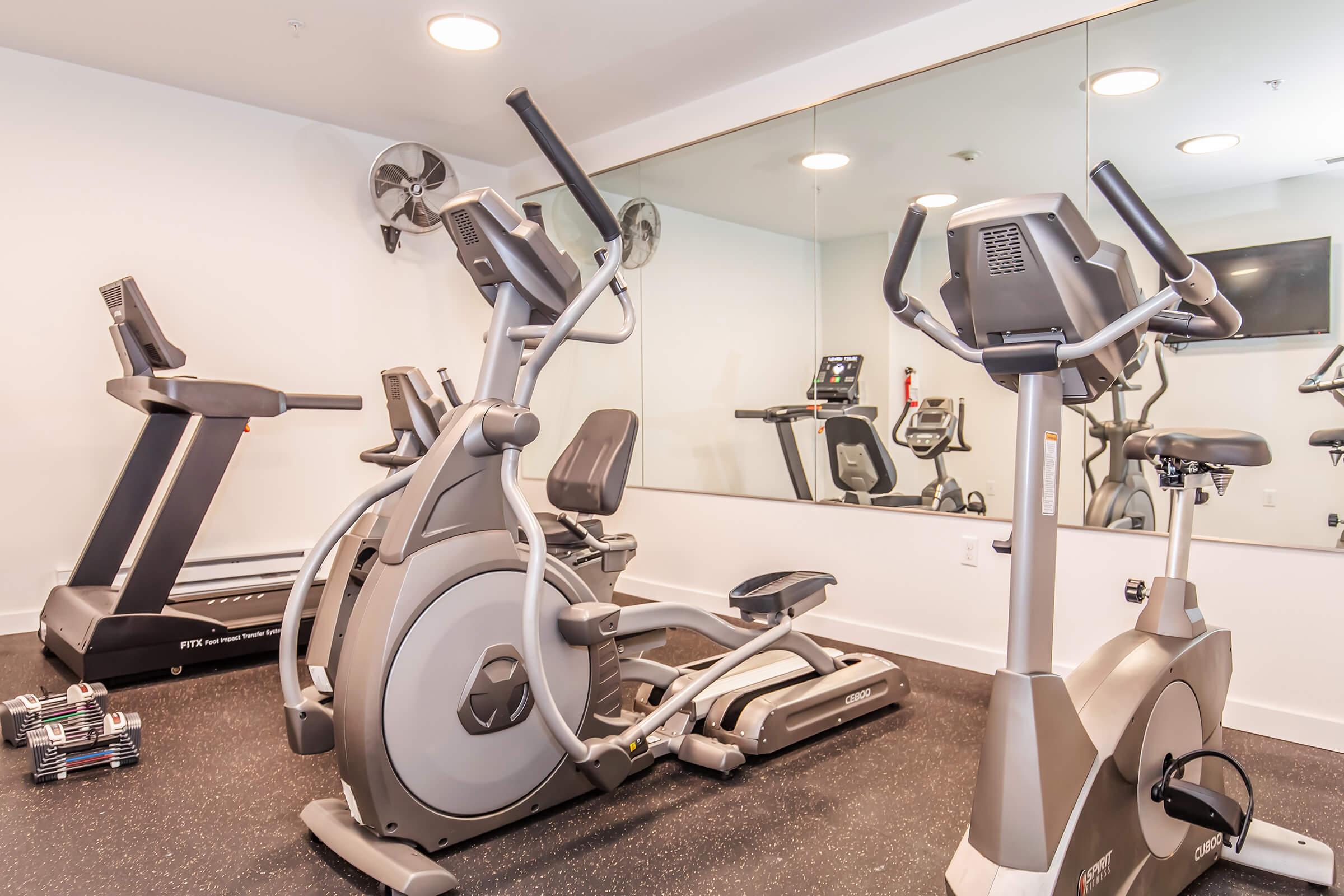 A modern gym featuring an elliptical machine, a treadmill, and a stationary bike, all set on a black rubber floor. There are mirrors on one wall, and a flat-screen TV is mounted in the corner. The space is well-lit and neatly organized, suggesting a comfortable environment for exercise.