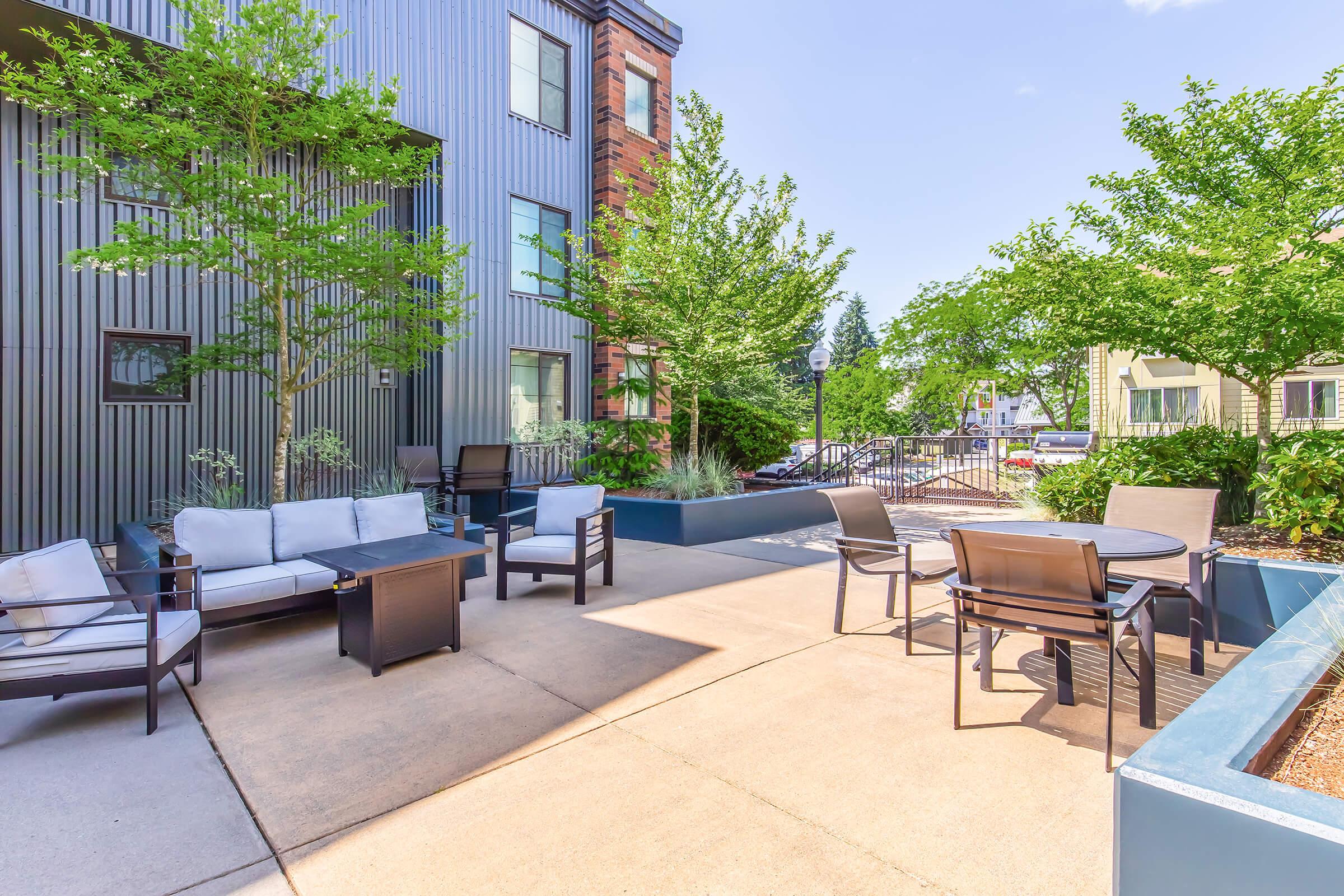 An outdoor patio area featuring modern seating with gray cushions, a coffee table, and a dining table with chairs. Lush green trees and shrubs surround the space, creating a relaxing atmosphere. The background shows a contemporary building with a mix of materials and finishes.