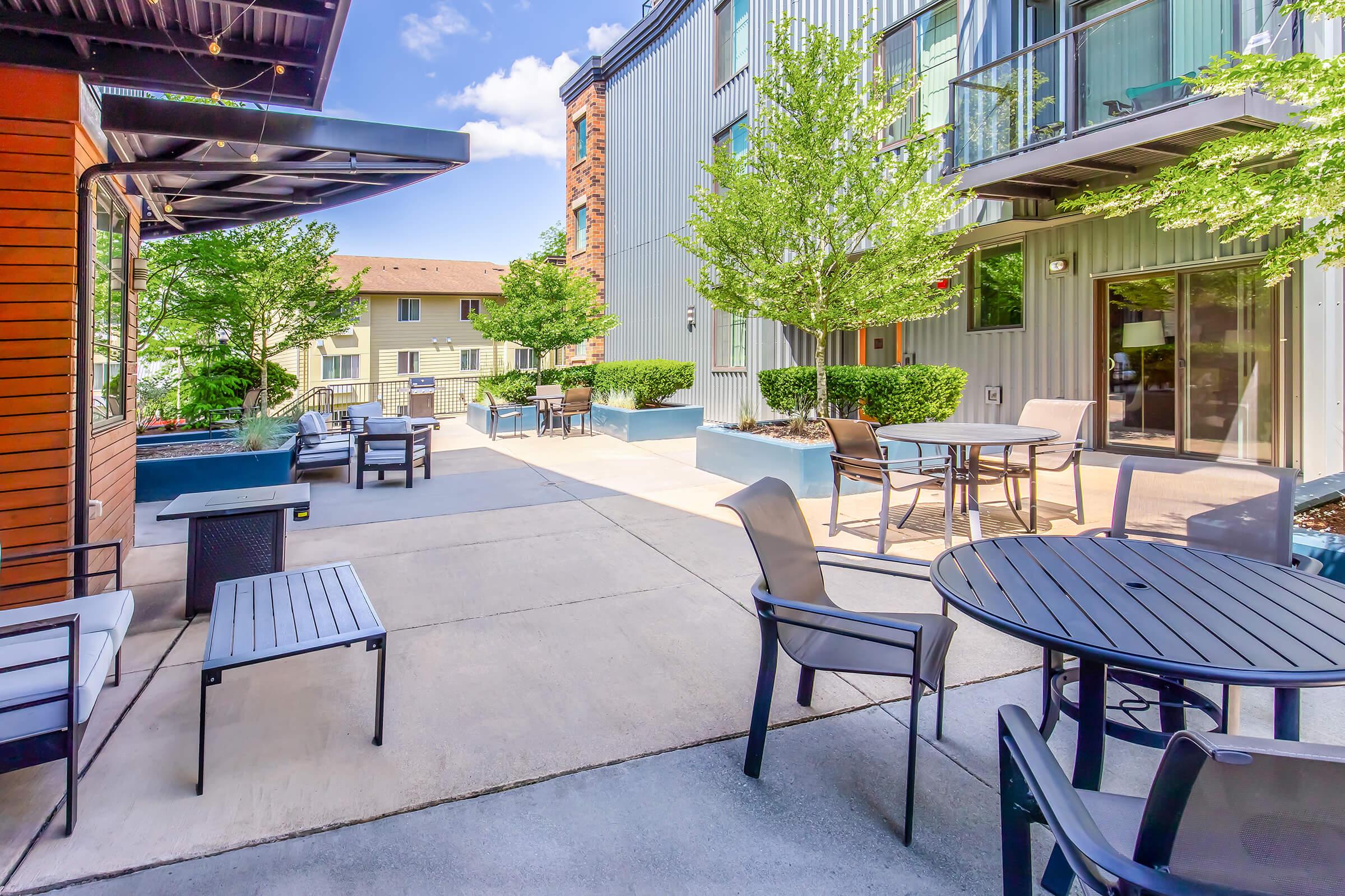 Outdoor space featuring modern furniture, including tables and chairs, surrounded by greenery and buildings. The area is well-lit with a clear blue sky, creating a welcoming atmosphere for relaxation or social gatherings.