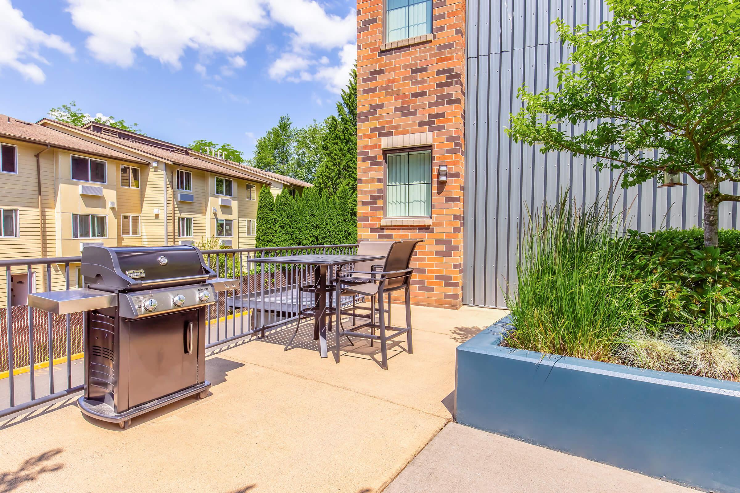 A modern grill next to a dining table and chairs on a patio, surrounded by greenery and a low decorative planter. In the background, there is a building with a brick facade and other apartment units visible under a clear blue sky.