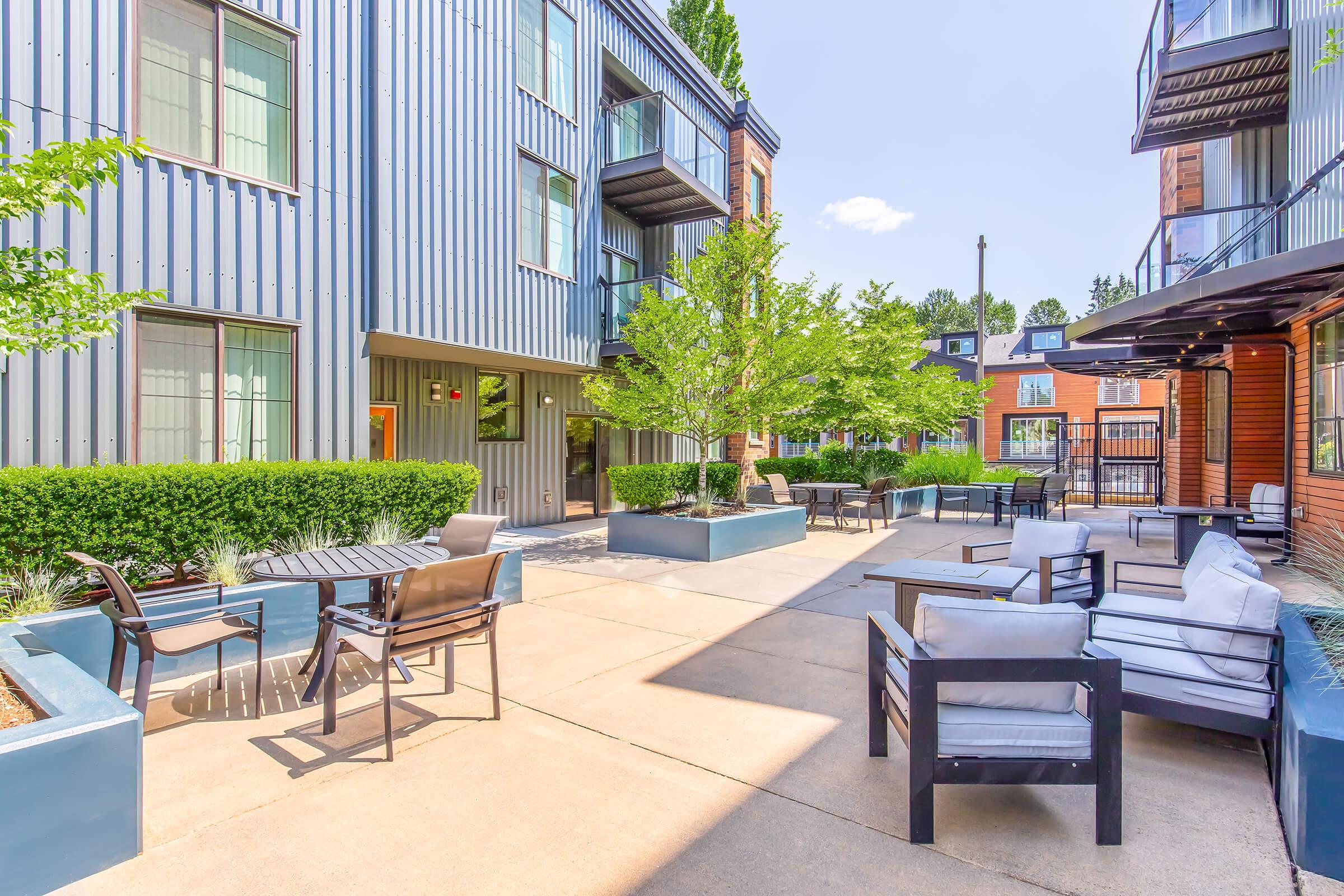 Outdoor patio area featuring modern furniture, including tables and chairs, surrounded by lush greenery and shrubs. The setting includes a mix of building styles with corrugated metal and wood accents, creating a welcoming environment for relaxation or social gatherings. Clear blue sky overhead.