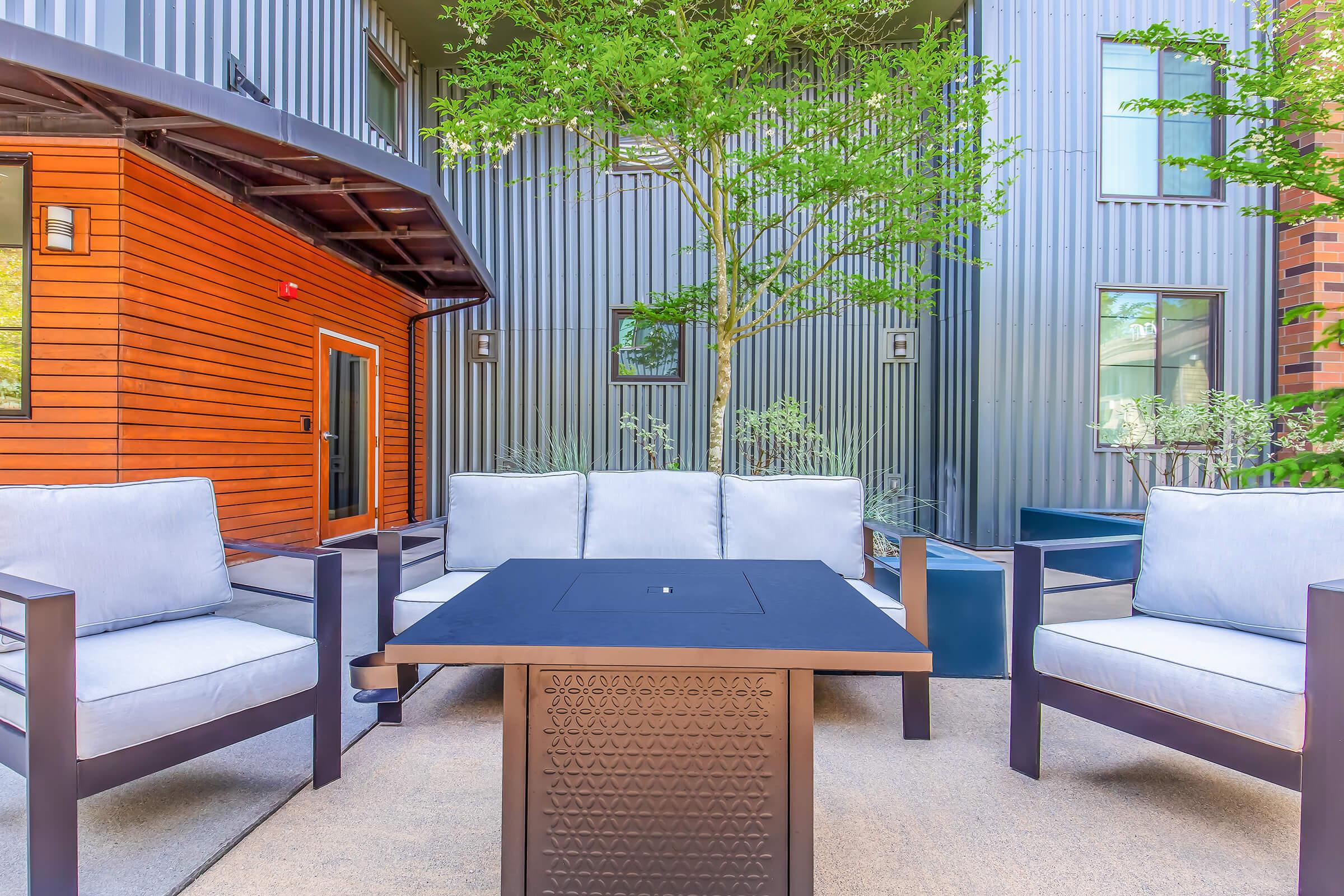 A cozy outdoor seating area featuring modern gray couches and a central fire pit table, surrounded by greenery and contemporary buildings with textured metal siding and wooden accents. Ideal for relaxation or gatherings in a stylish urban setting.