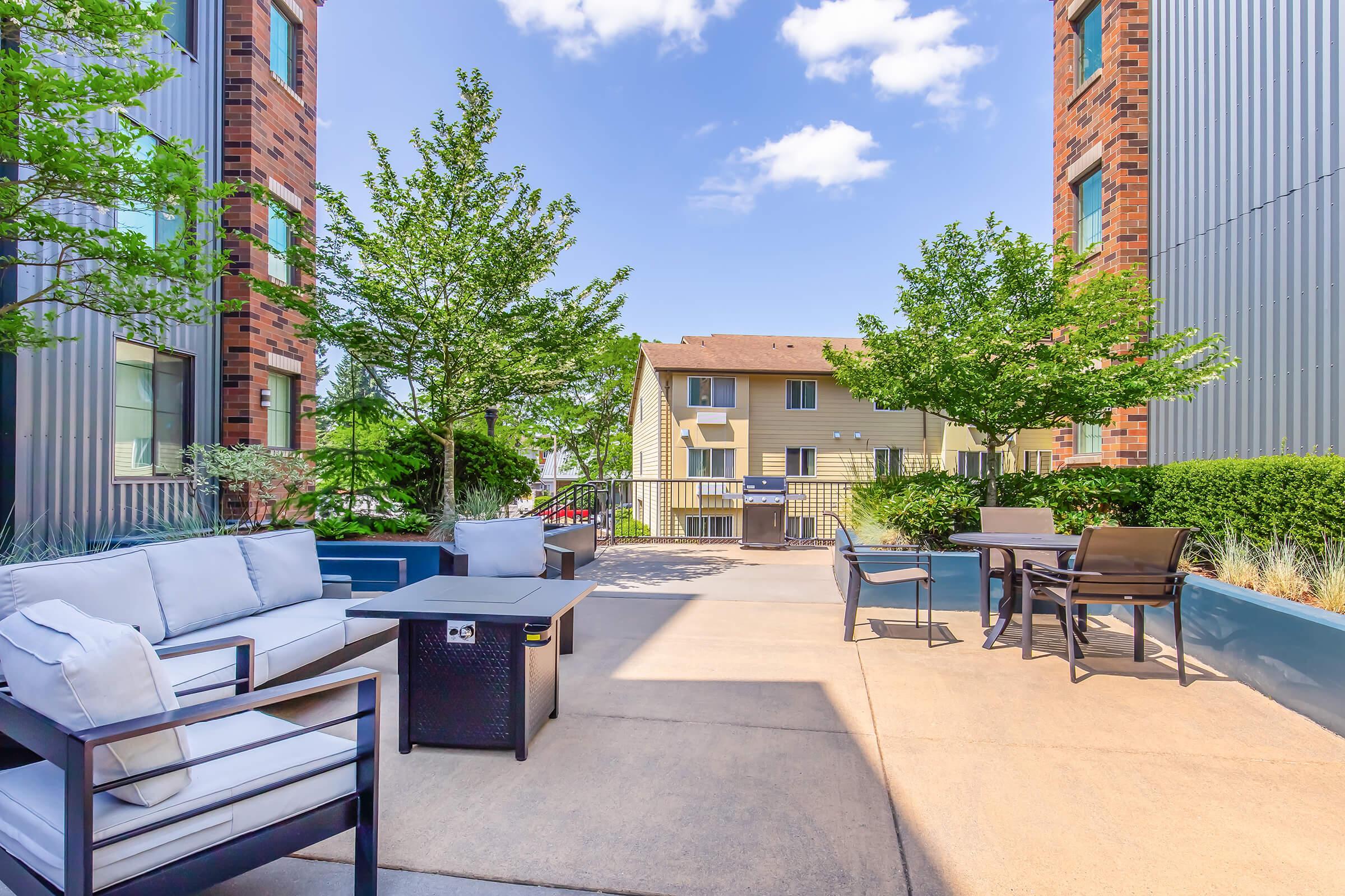 Outdoor patio area featuring modern seating with gray cushions, a small black coffee table, and surrounded by green trees. The background includes several buildings, showcasing a sunny blue sky with a few clouds.
