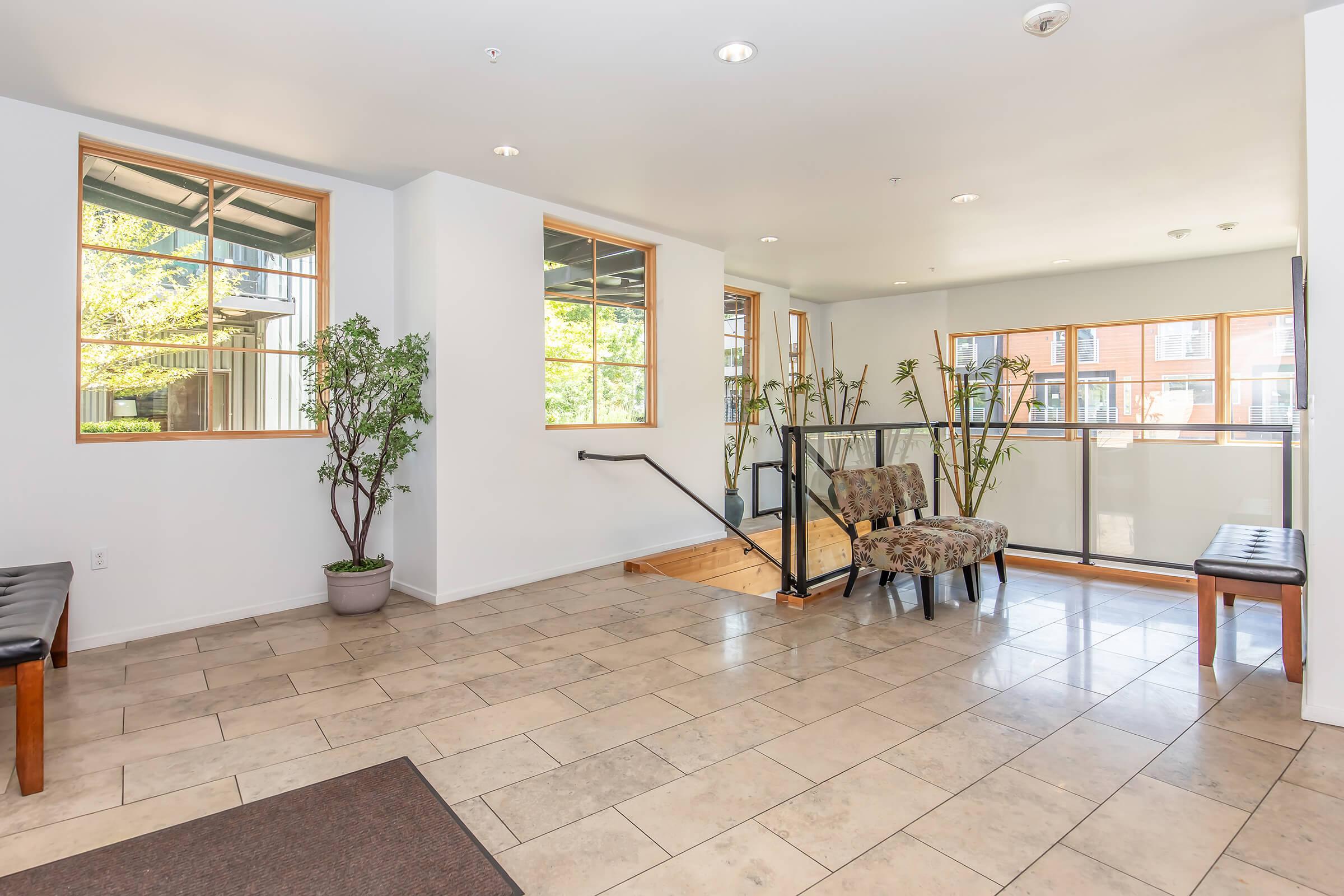 Interior view of a bright, modern lobby area featuring tiled flooring, large windows allowing natural light, potted plants, a cozy chair, and a wooden bench. There is a staircase leading to another level, enhancing the spacious atmosphere.