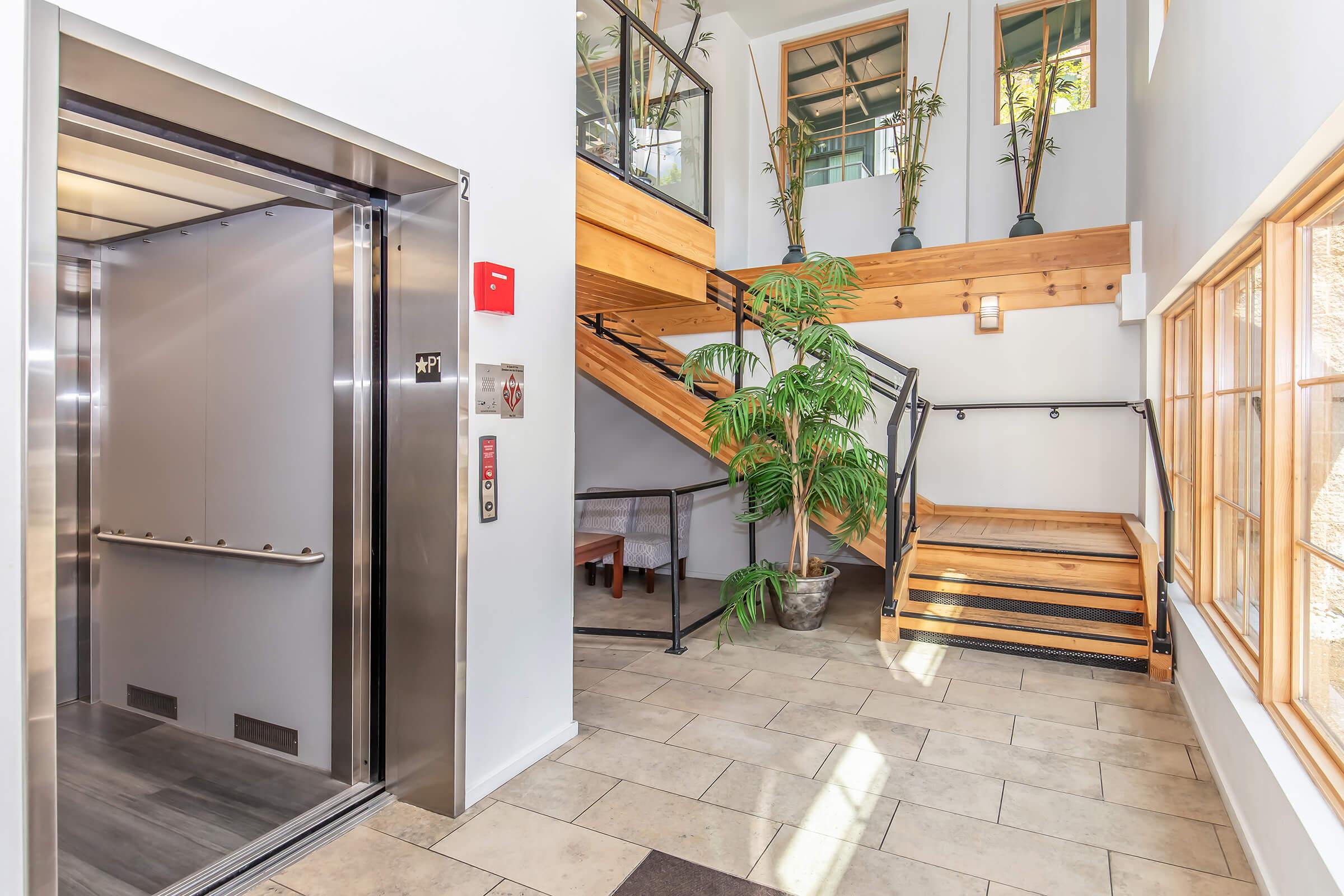 Modern interior of a multi-level building featuring an elevator, wooden stairs, and a lush plant. Natural light floods the space through large windows, highlighting the warm tones of the wooden accents. An inviting atmosphere is created by the combination of contemporary architecture and greenery.