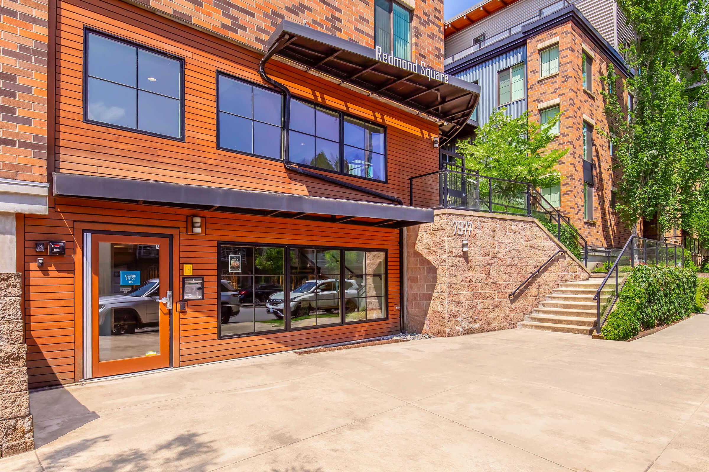 Exterior of a modern brick building featuring large windows and a prominent entrance with a glass door. The building is situated next to a stone pathway with stairs leading up to a landscaped area. Green trees can be seen nearby, enhancing the urban setting.