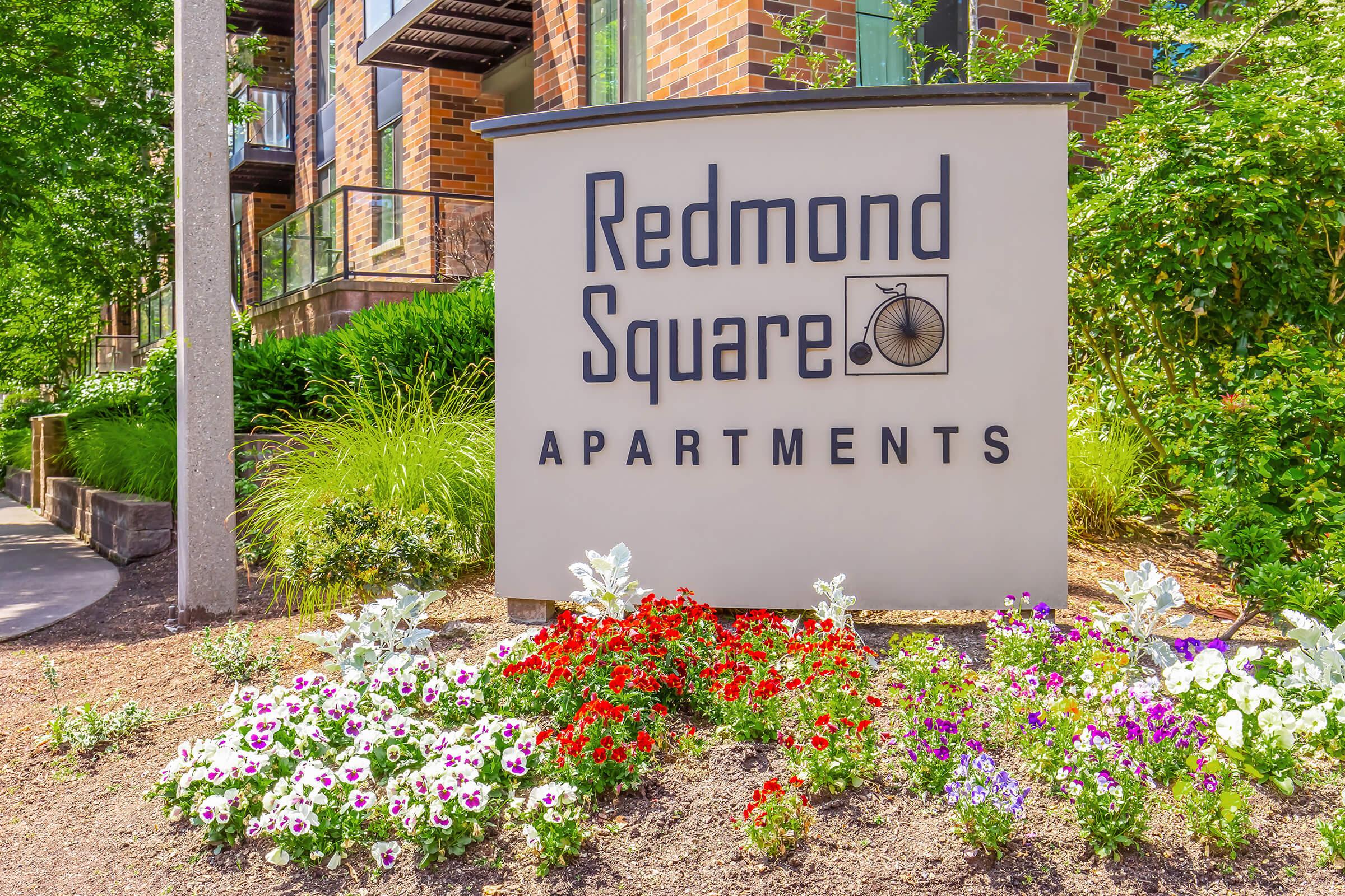 Sign for Redmond Square Apartments featuring the name prominently displayed. The sign is surrounded by colorful flowers, including petunias, and lush green foliage, set against a backdrop of a modern brick building. The environment appears well-maintained and inviting.