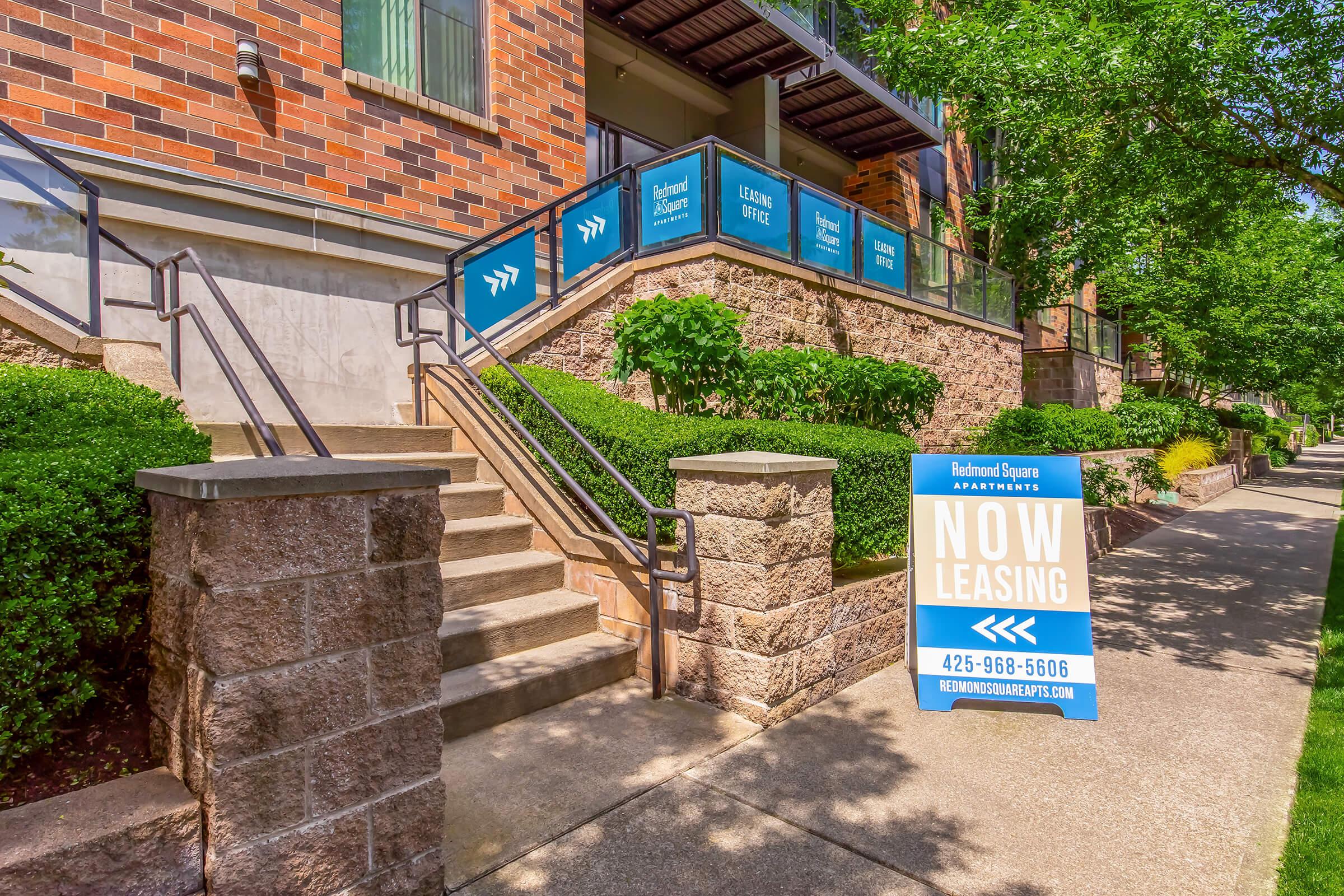 Stairs leading to an apartment complex with a modern design. Nearby, there are signs advertising leasing opportunities. The building features large windows and is surrounded by well-kept greenery, including hedges and trees, creating an inviting atmosphere. A sidewalk runs alongside the property.