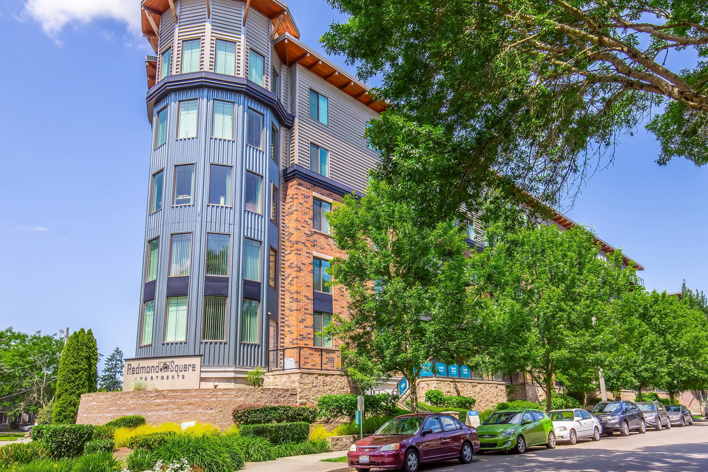 Modern apartment building featuring a mix of brick and glass architecture. The structure has multiple stories with a distinct tower design. Lush trees and well-maintained landscaping surround the building, while several parked cars line the street in front. Bright blue sky adds to the inviting atmosphere.
