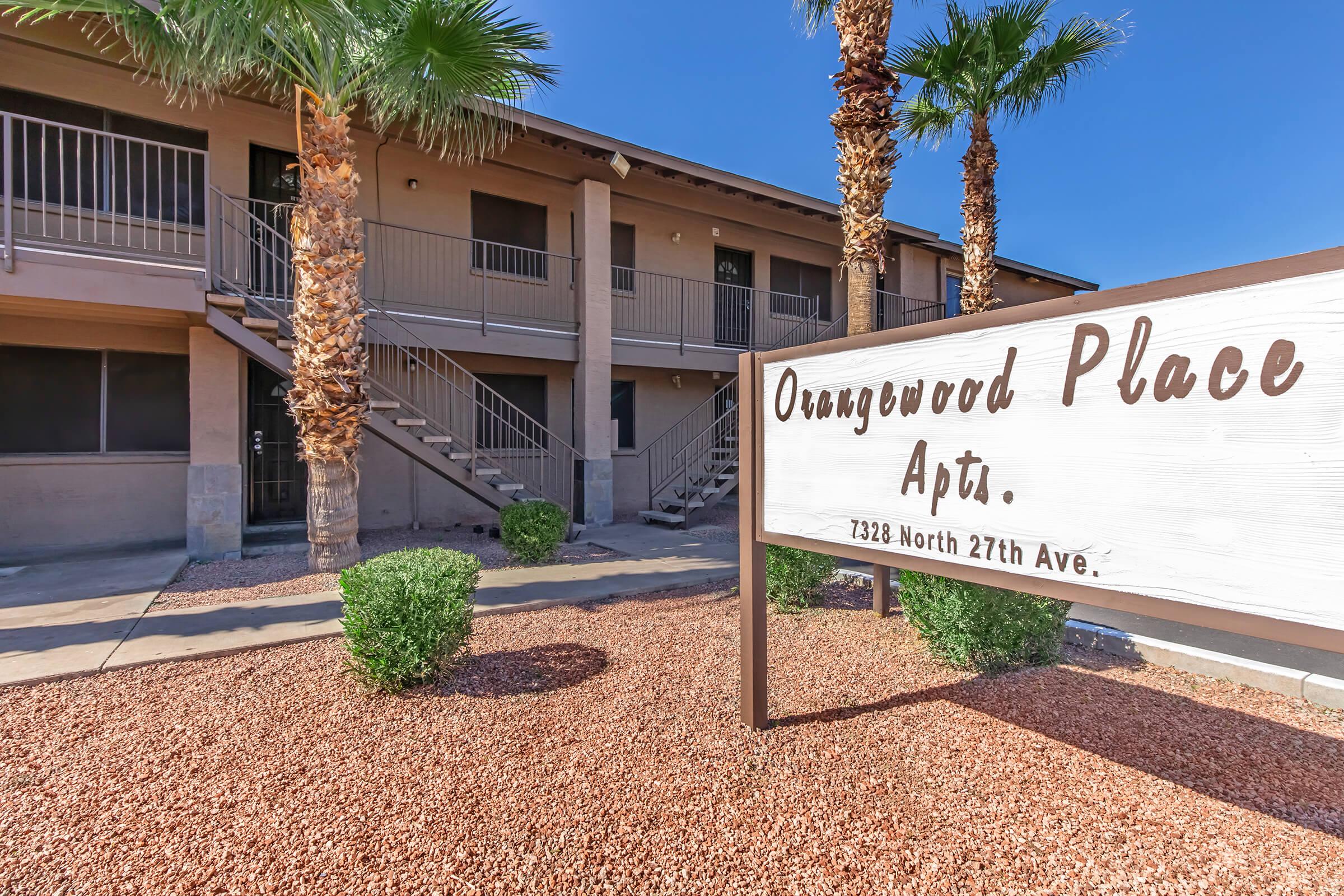 Sign for "Orangewood Place Apartments" with a landscaped area featuring small bushes and palm trees. The building is a two-story structure with staircases leading to the second floor, set against a clear blue sky.
