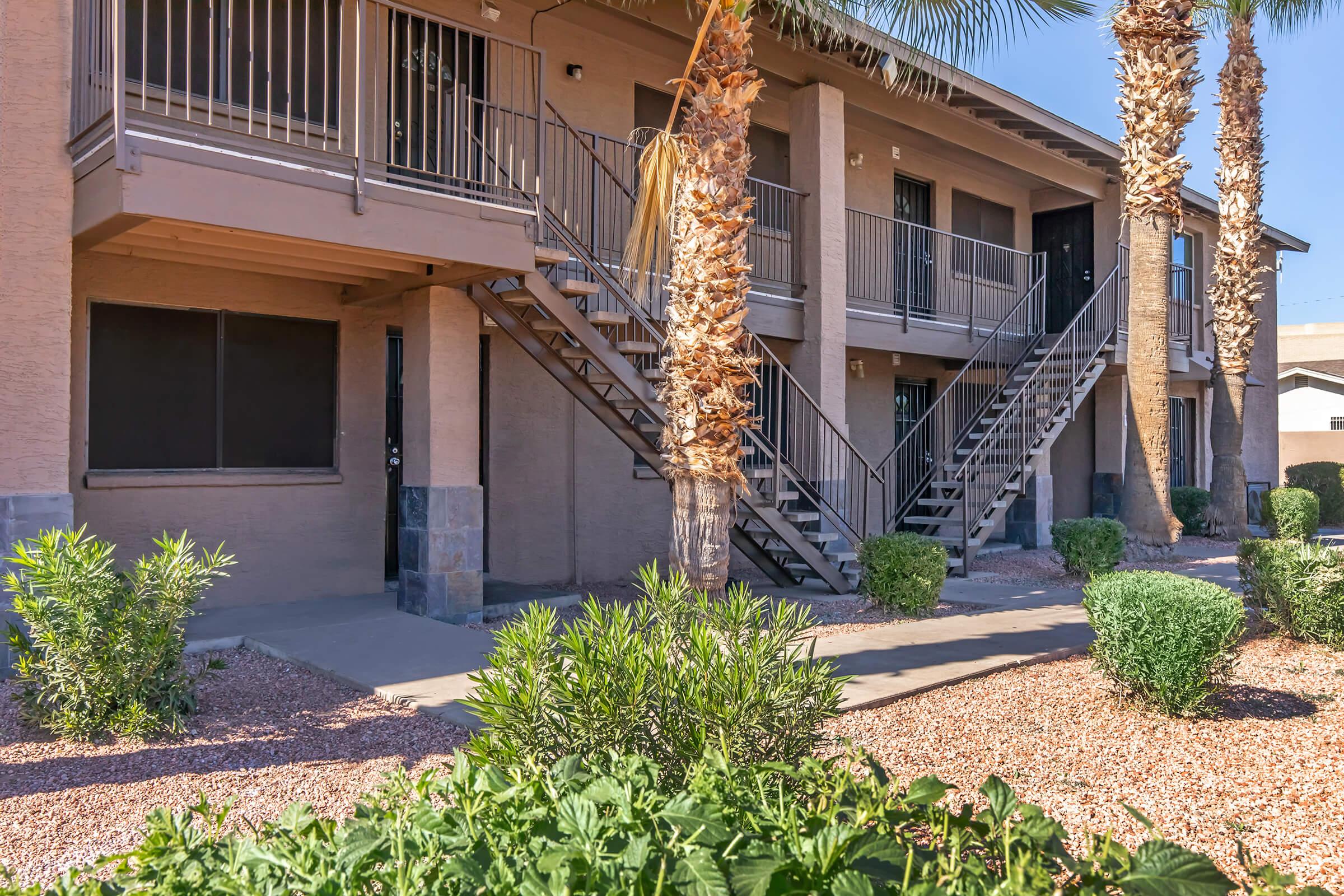 A view of an apartment complex featuring two levels with metal staircases, surrounded by landscaped greenery and pebbled pathways. Palm trees add to the tropical ambiance, while the clear blue sky complements the scene.