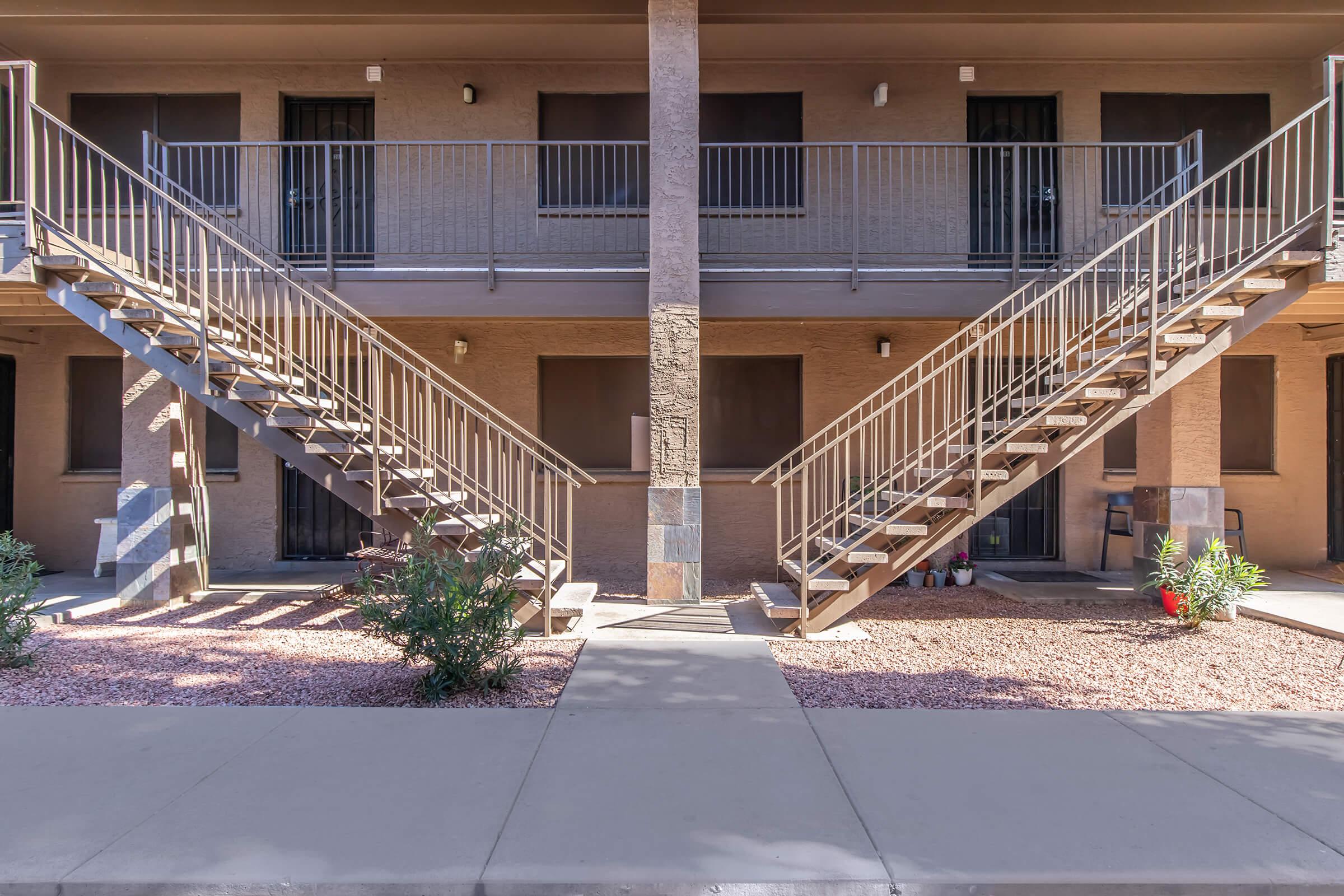 Two sets of metal staircases lead to upper-level entrances of a brown, multi-unit building. The staircases are flanked by a pathway lined with small shrubs and gravel. The building features a simple, modern design with doors that have security bars and no visible windows. The area appears neat and well-maintained.