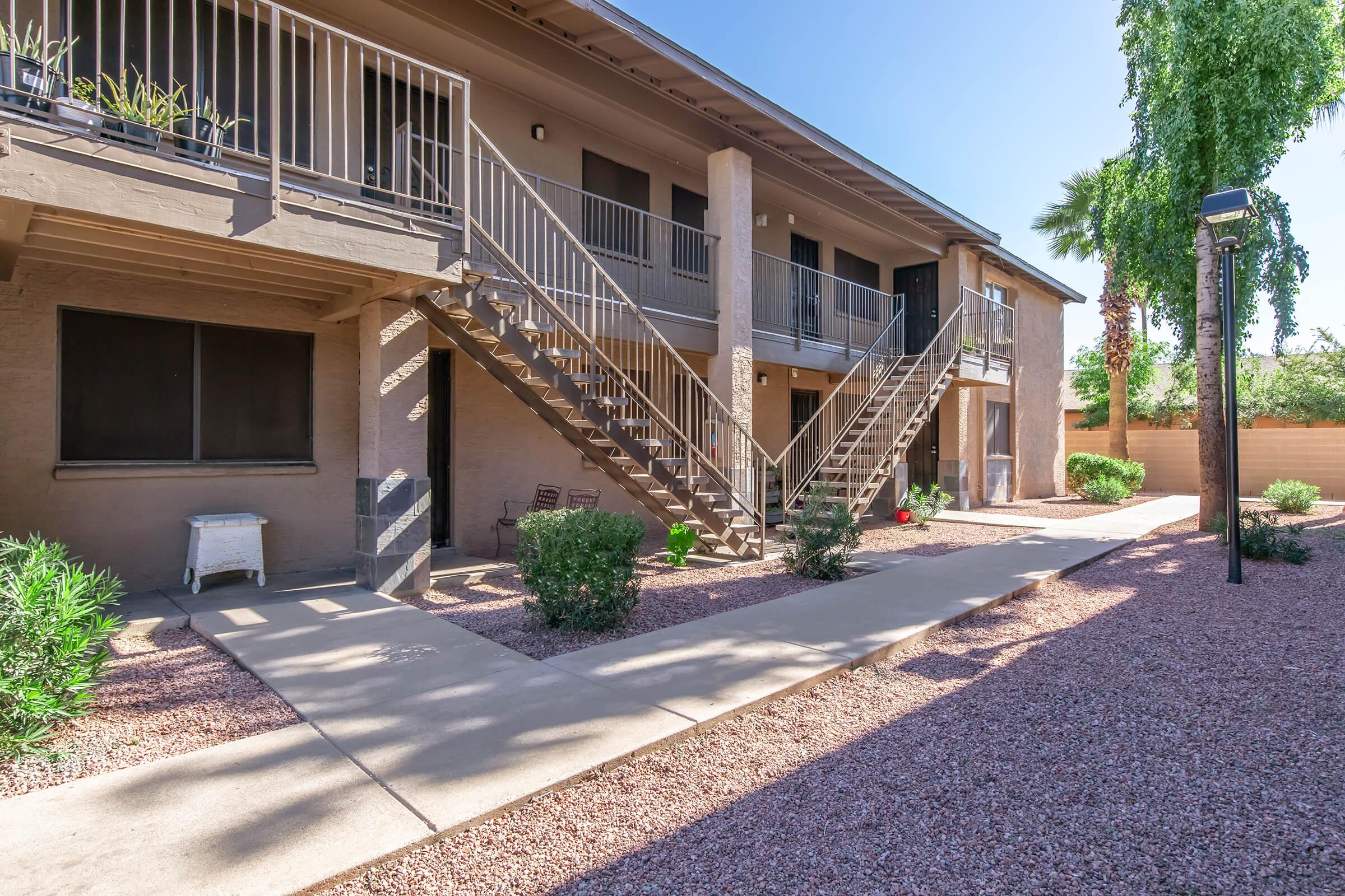 A view of a two-story apartment building featuring outdoor staircases, with a landscaped path flanked by small bushes and gravel. The entrance has a few chairs and potted plants, and the setting is under clear blue skies, showcasing a well-maintained outdoor area.