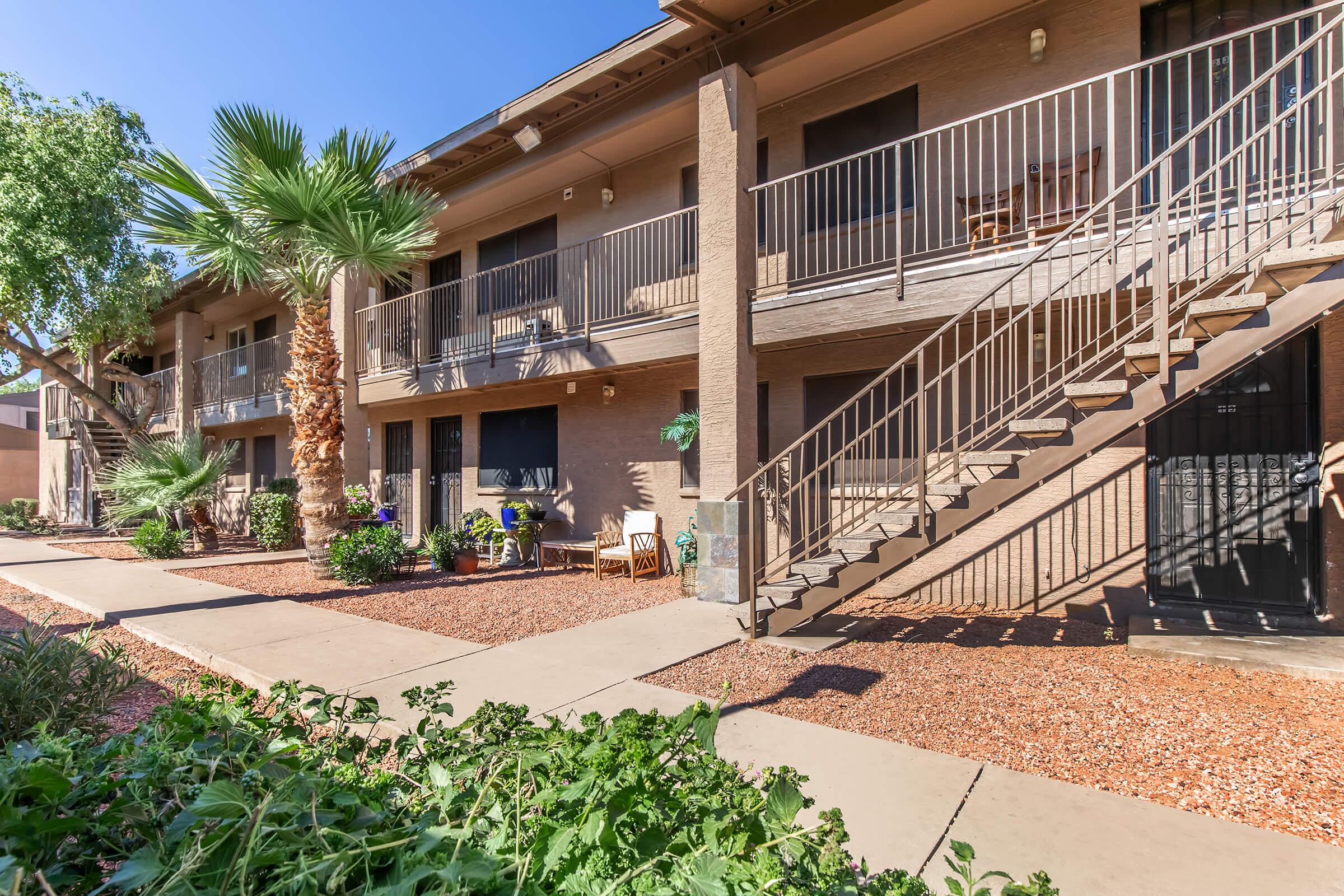 A view of a multi-unit apartment building featuring palm trees, outdoor stairs, and a landscaped pathway with plants. The facade is beige with balconies, and chairs are set outside some units. The scene is bright and sunny, highlighting a pleasant residential area.