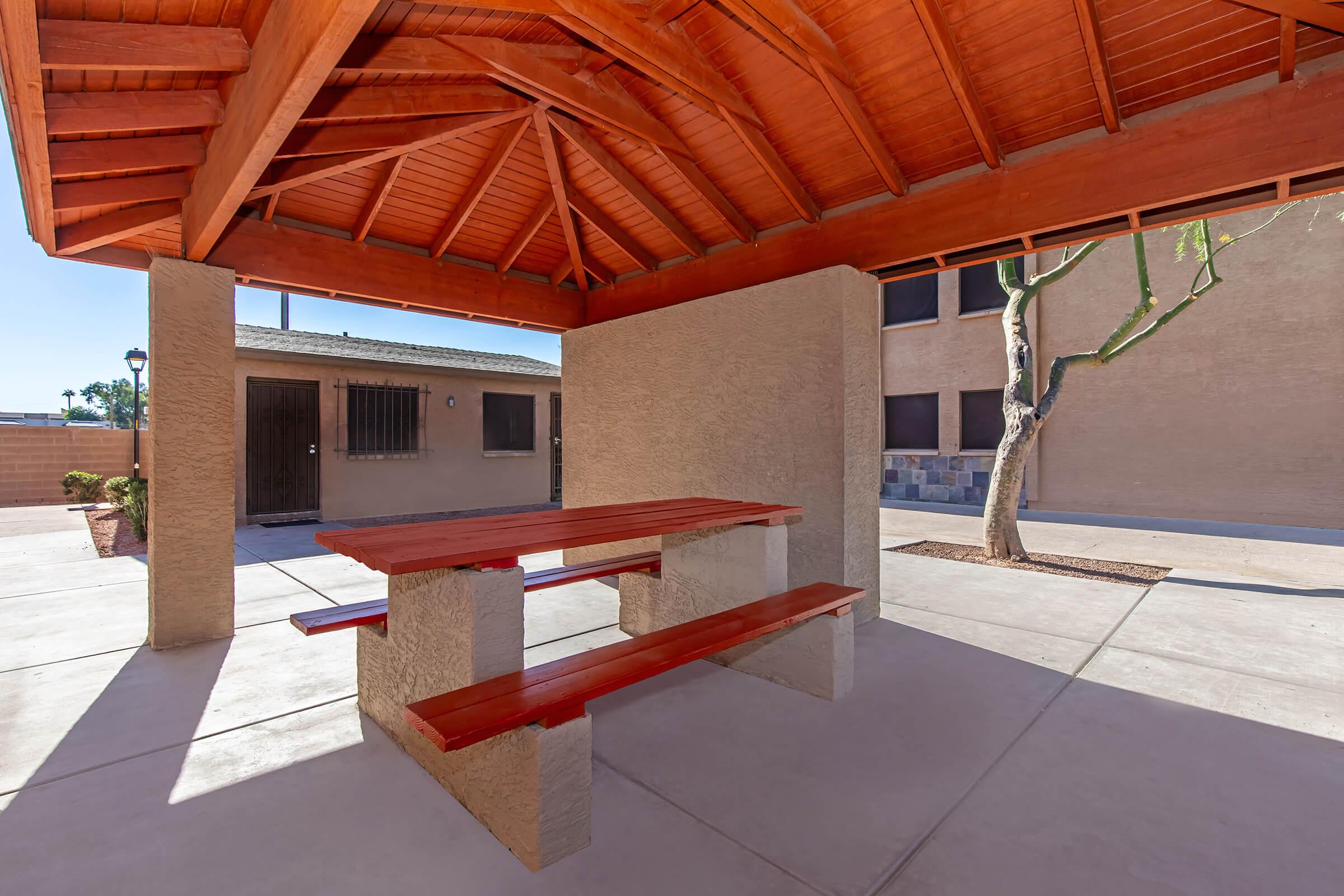 A shaded outdoor space featuring a wooden picnic table with benches, under a wooden structure with a slanted roof. The area is surrounded by beige walls and includes a nearby building with windows, offering a clean and inviting atmosphere for gatherings or relaxation.