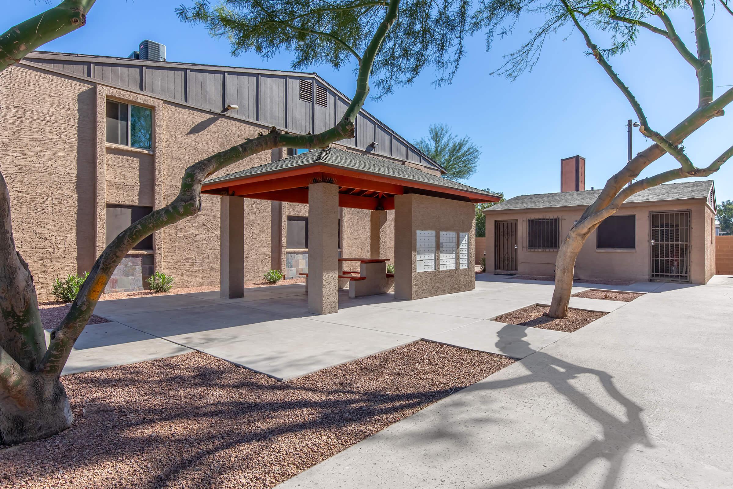 A shaded gazebo in a courtyard surrounded by trees, with a concrete pathway and building facades in the background. The space is well-lit and designed for outdoor use, featuring areas for seating and relaxation.