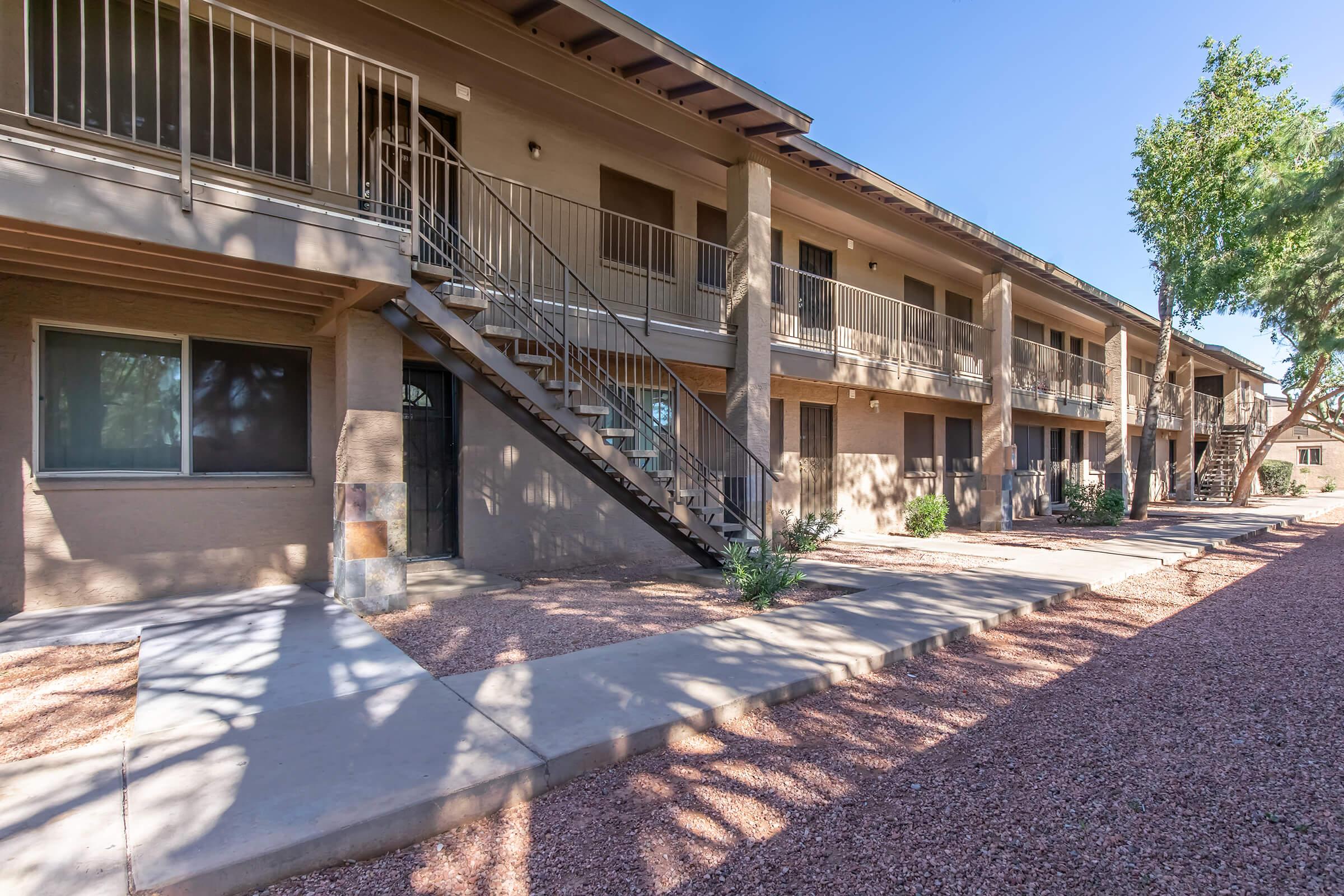 A view of a two-story apartment complex with a staircase leading to the second floor. The building features balconies and walkways surrounded by small plants and gravel landscaping. Trees provide some shade, and the sky is clear and sunny.