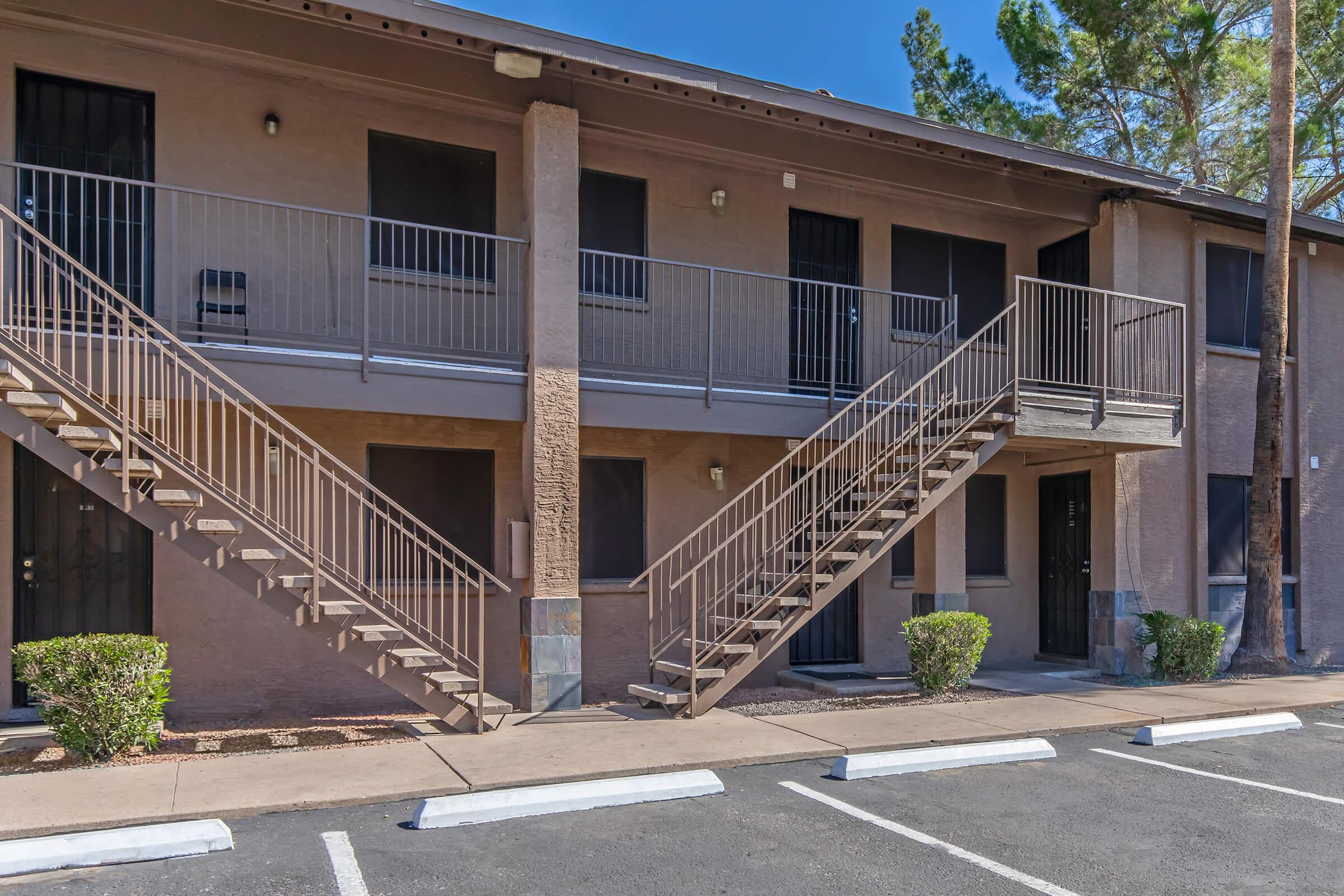 Two-story apartment building featuring a sandy brown exterior, with metal staircases leading to second-floor entrances. The ground level has two doors, and the area is landscaped with small shrubs. Parking spaces are visible in front, with a clear blue sky above.