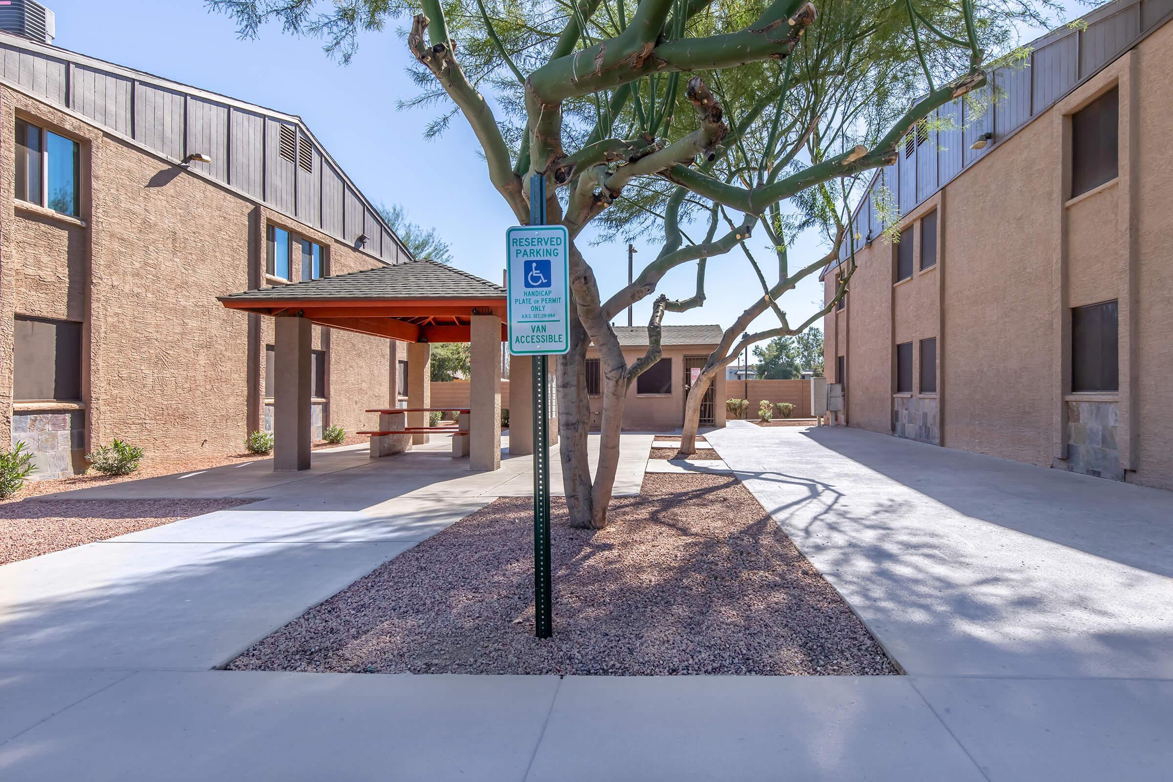A landscape view of a pathway between two buildings with a parking sign indicating reserved parking for individuals with disabilities. The scene includes a tree and a shaded gazebo area, all set in a well-maintained outdoor space, featuring gravel and concrete surfaces under a clear blue sky.