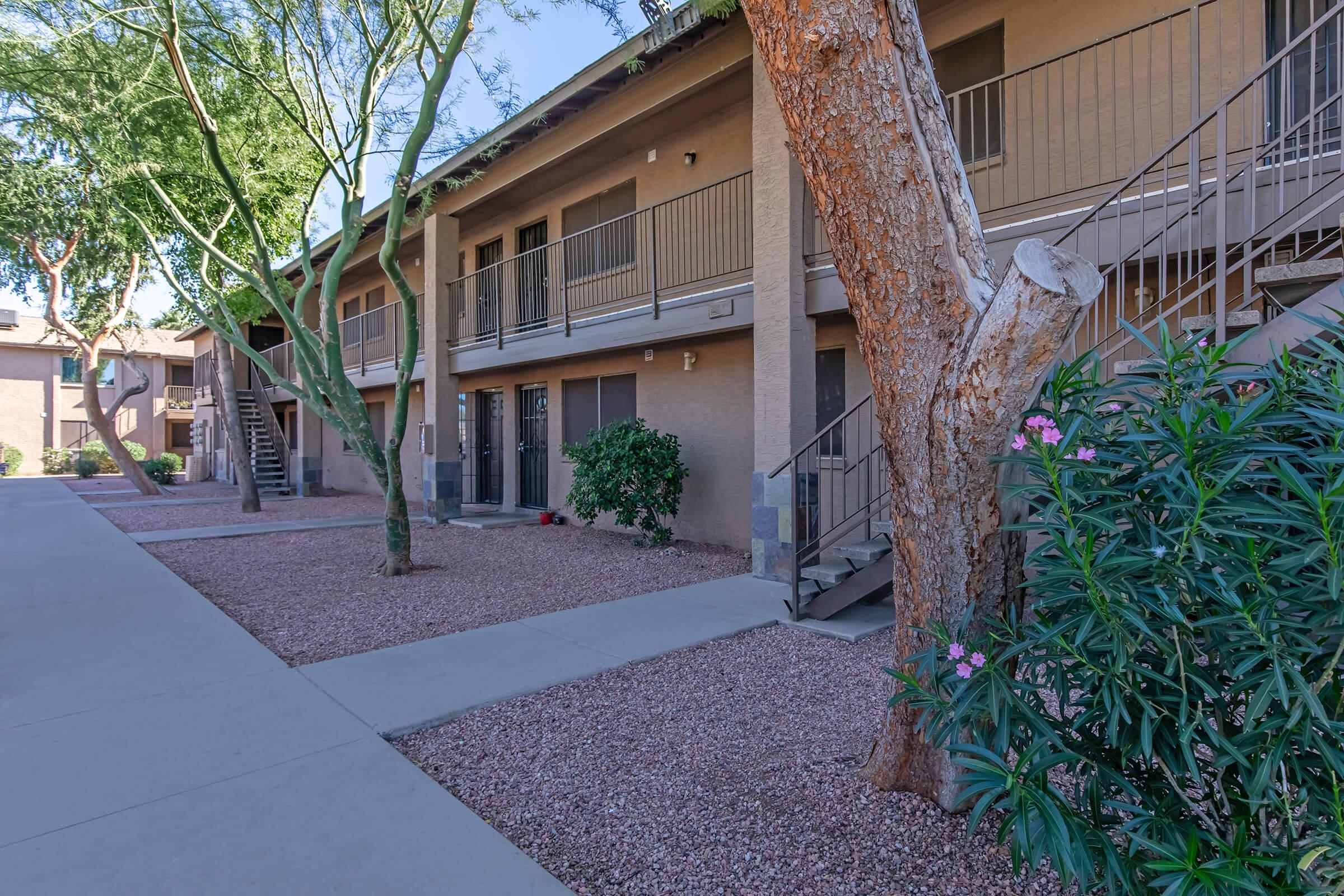 View of a residential apartment complex featuring two-story buildings with balconies, surrounded by landscaped pathways, trees, and decorative shrubs. The area is well-maintained, with gravel ground cover and a sunny atmosphere, highlighting the inviting outdoor space.