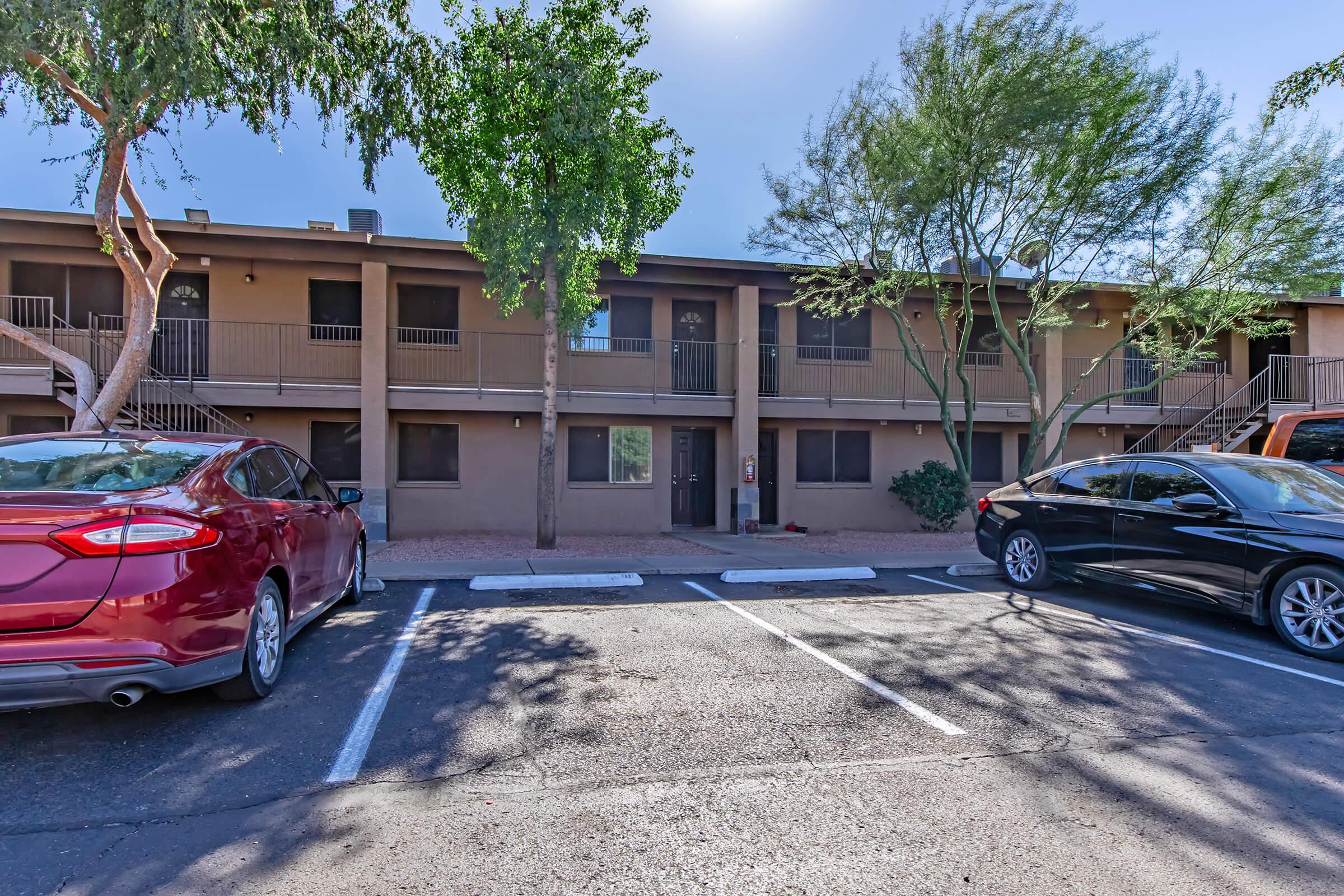 A view of an apartment building with two stories, featuring several trees and parked cars in front. The building has balconies and entrances along the ground floor. The parking lot has designated spaces with some shade from the trees. The scene is bright and sunny, typical of a warm climate.