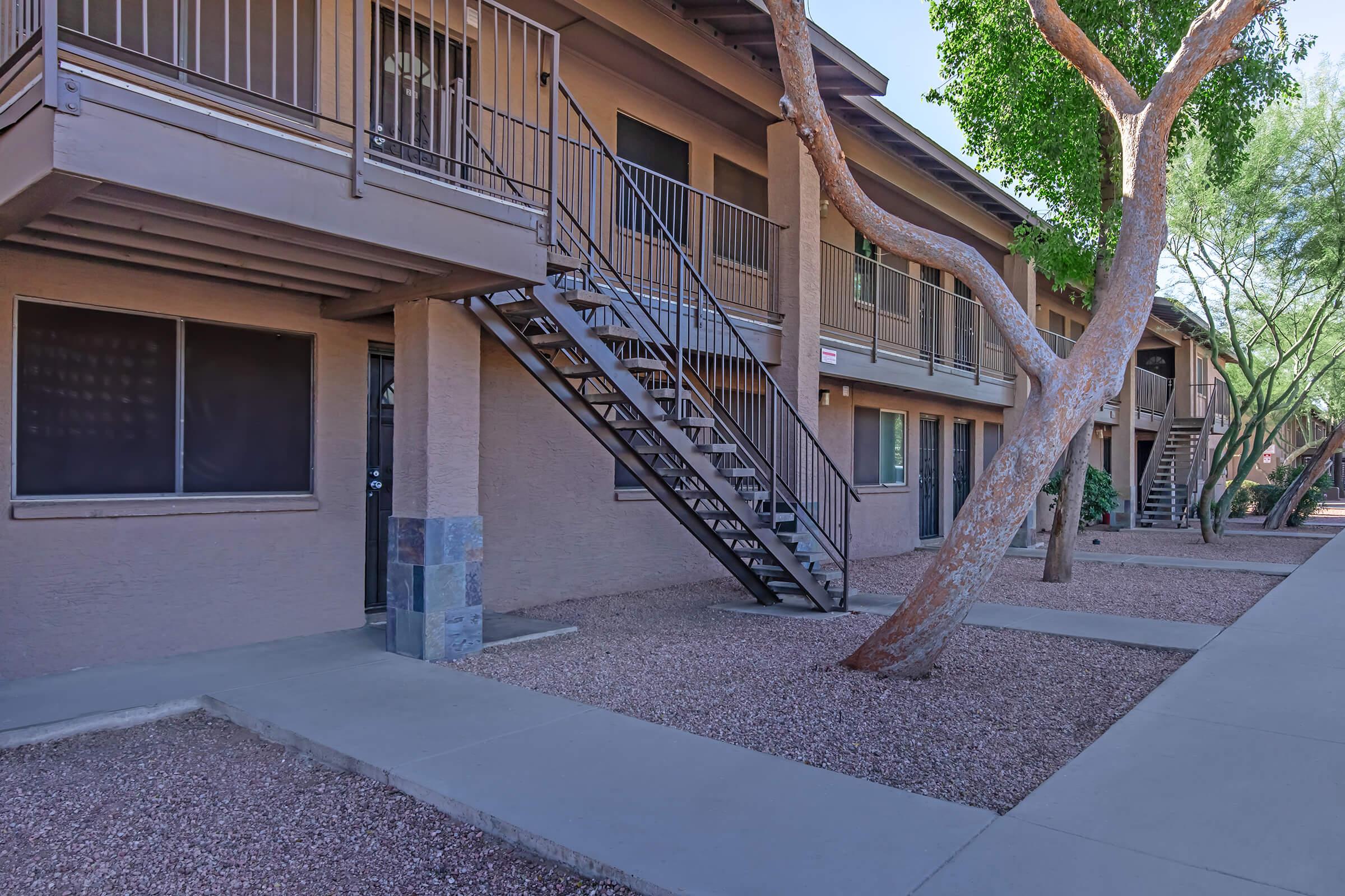 Exterior view of a two-story apartment building with multiple units. A staircase leads to the upper floor. The building features a neutral color palette, large windows, and a gravel landscape. Trees and shrubs are visible alongside the walkway, providing greenery to the setting.