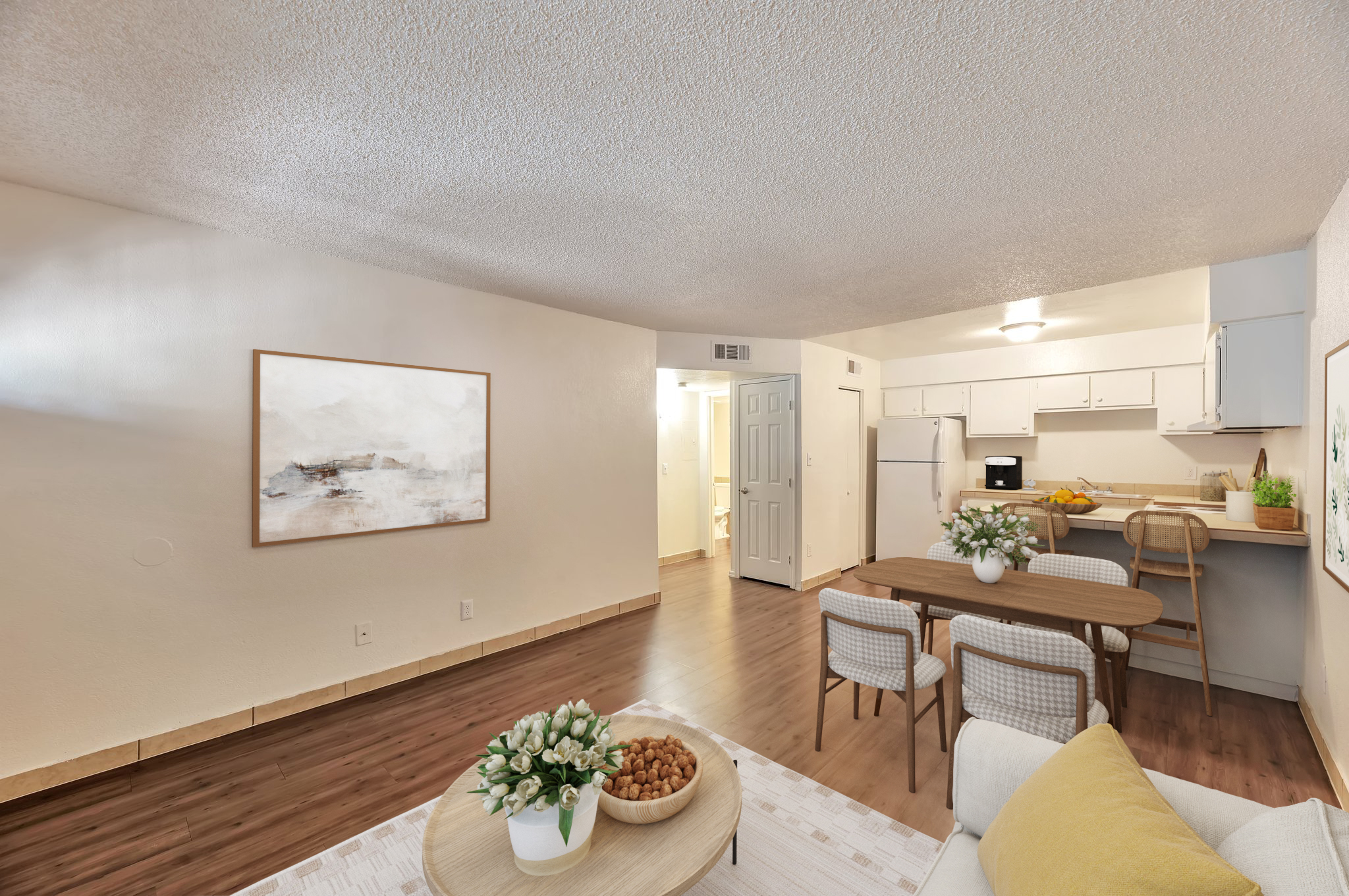 A cozy living space featuring a dining table with chairs, a comfortable sofa, and a decorative bowl on a coffee table. The light-colored walls and wood flooring create an inviting atmosphere. The kitchen is visible in the background, with white cabinets and a door leading to a bathroom.