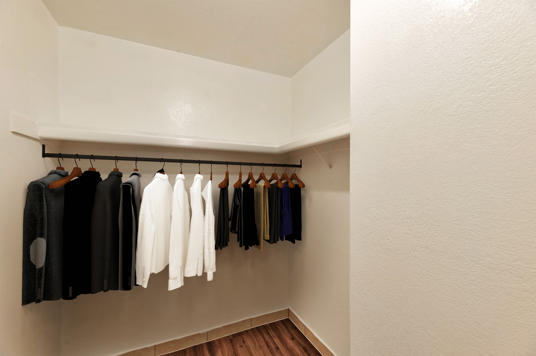 A minimalist closet featuring a white wall and a wooden floor. It contains a rod with several neatly hung garments, including a mix of black, white, and colored shirts. The empty space emphasizes the organized layout, with a bright and clean aesthetic.
