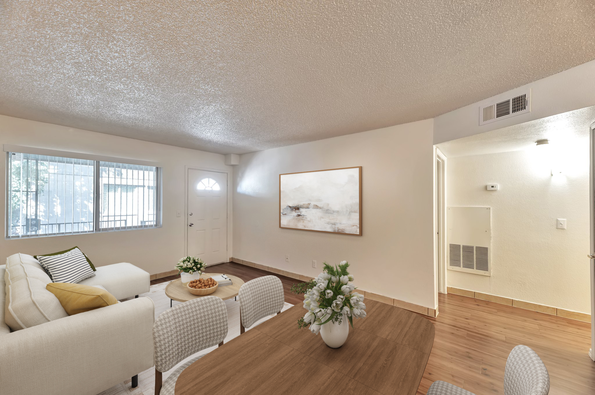 A cozy living space featuring a beige sofa with striped cushions, a wooden dining table set with flowers, light-colored walls, and a large window allowing natural light. The room has hardwood floors and a simple decorative piece on the wall, creating a warm and inviting atmosphere.
