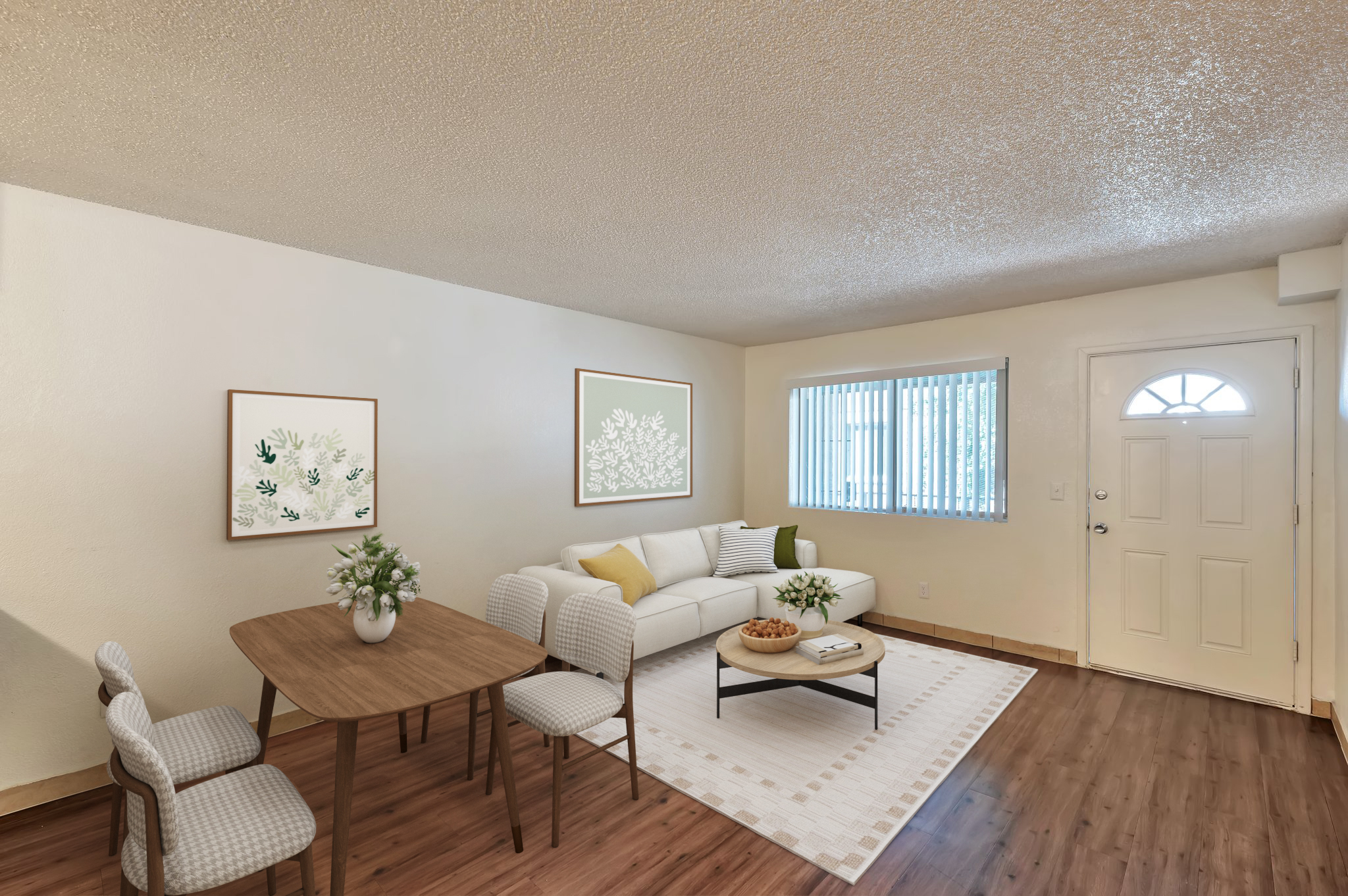 A cozy living room featuring a white sofa with decorative pillows, a wooden dining table with chairs, and framed floral artwork on the walls. Natural light enters through a window by the front door, enhancing the warm, inviting atmosphere of the space.
