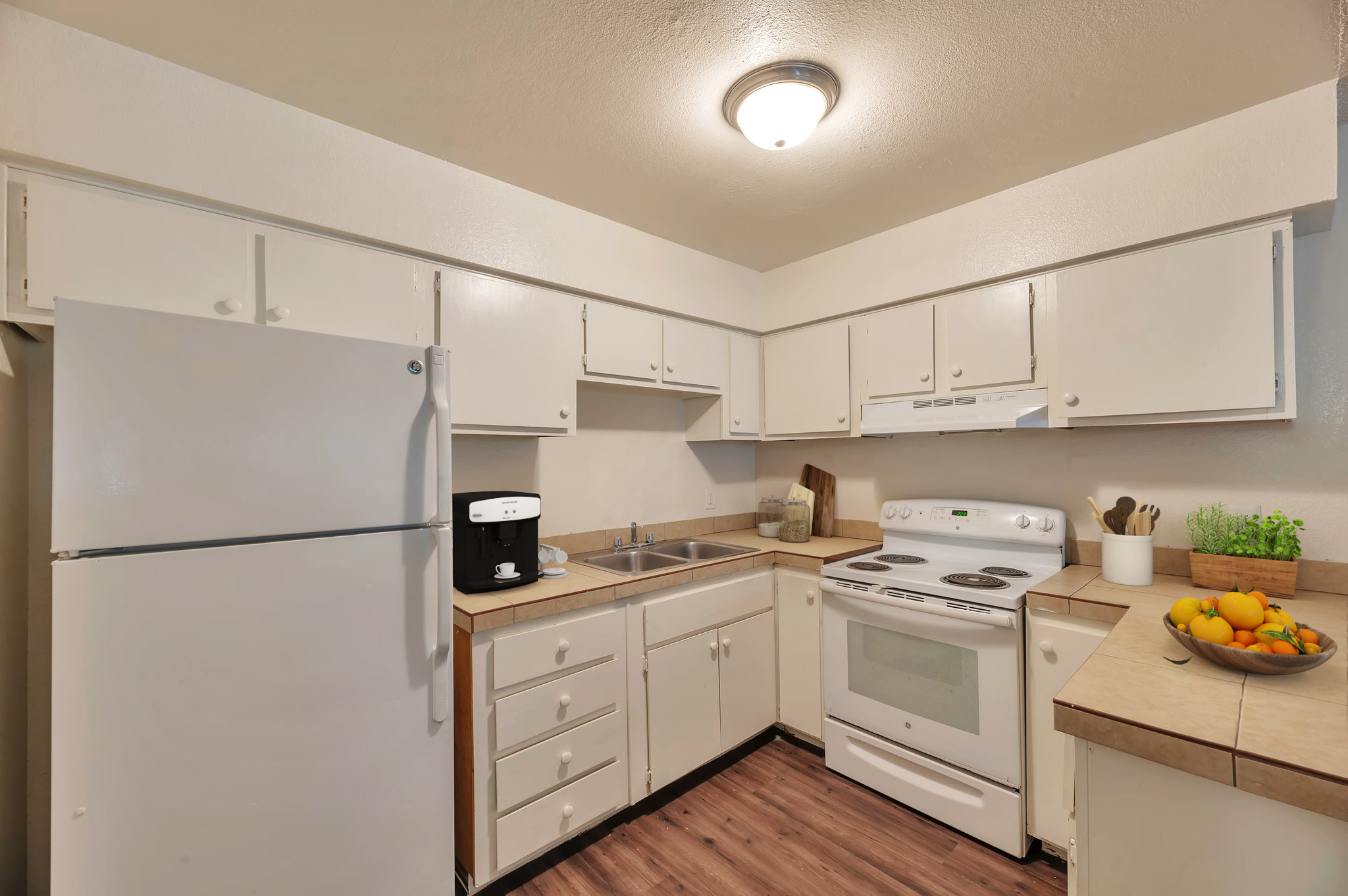 A clean and bright kitchen featuring white cabinets, a white refrigerator, a stove, and a sink. There's a countertop with a coffee maker, utensils, and a bowl of fresh fruits. The flooring is wood-like, and the room is well-lit with a ceiling light.