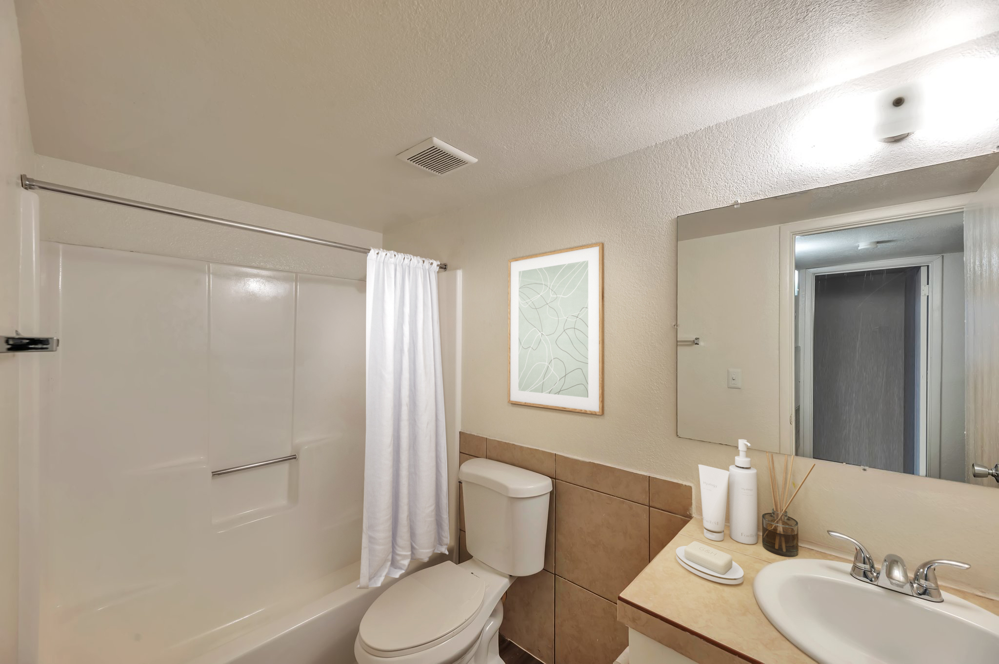 A bright and clean bathroom featuring a shower with a white curtain, a toilet, and a sink with toiletries. A large mirror reflects the space, and a decorative wall art piece adds elegance. Neutral tones dominate the tile and walls, creating a calming atmosphere.