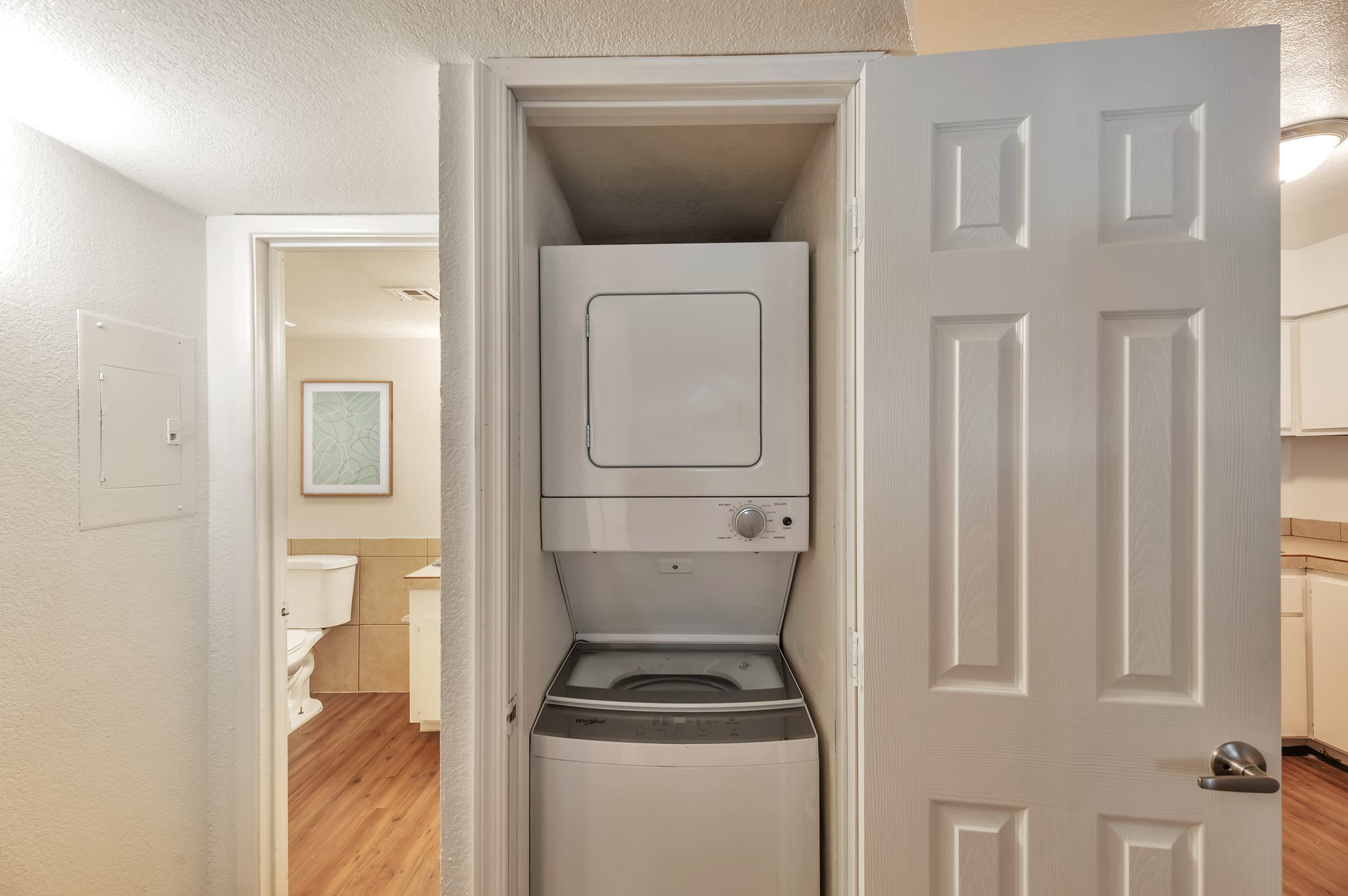 A stacked washer and dryer unit located in a small closet. The surrounding area is a well-lit hallway that leads to a bathroom. Light-colored walls and wooden flooring create a clean and tidy appearance. The closet doors are white and slightly ajar, showcasing the laundry appliances.