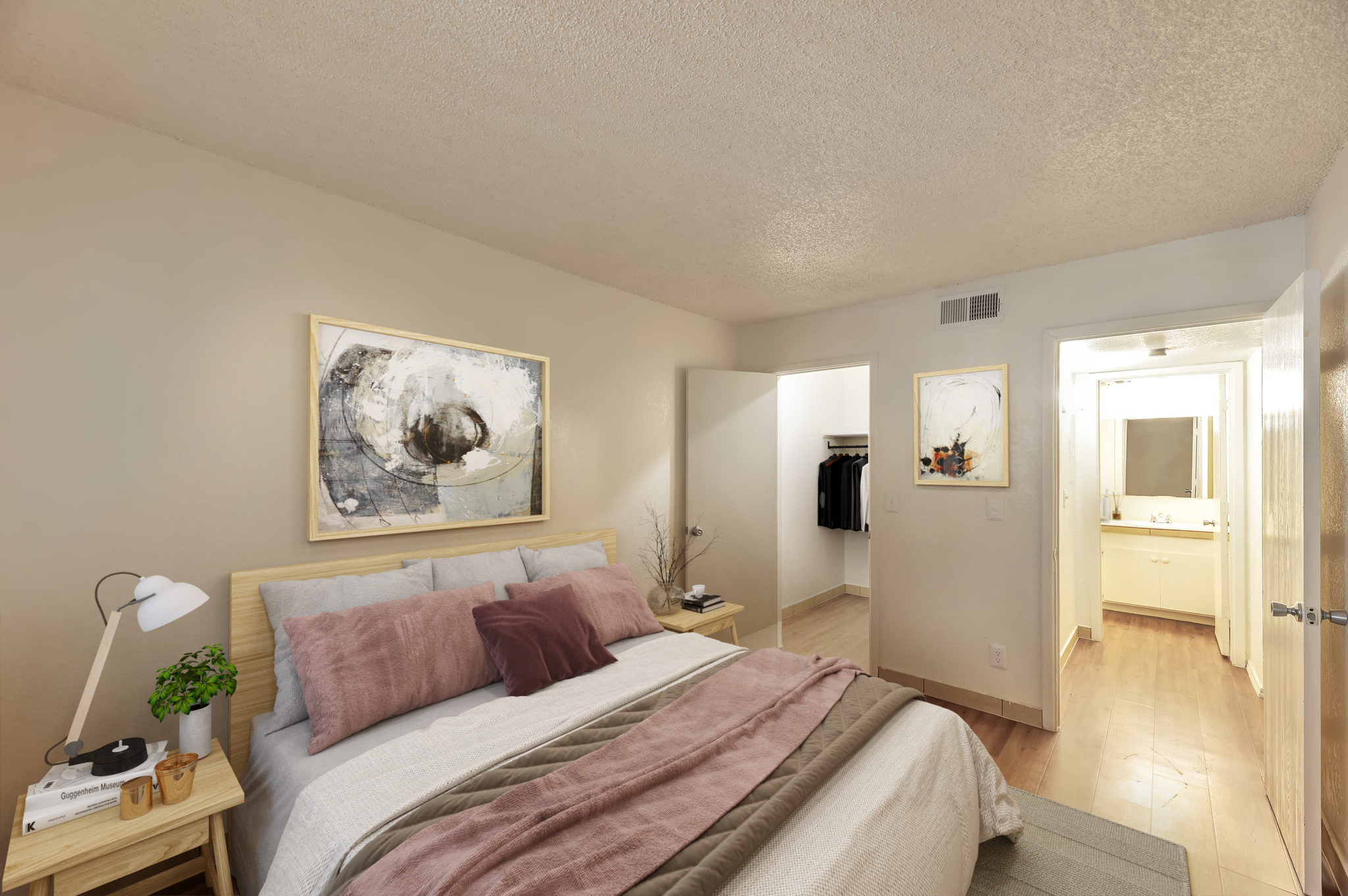 A cozy bedroom featuring a bed with gray and pink bedding, a bedside table with a lamp, and abstract wall art. A closet is visible in the background, along with a bathroom door that leads to a clean, well-lit bathroom. The room has light-colored walls and wooden flooring, creating a warm atmosphere.