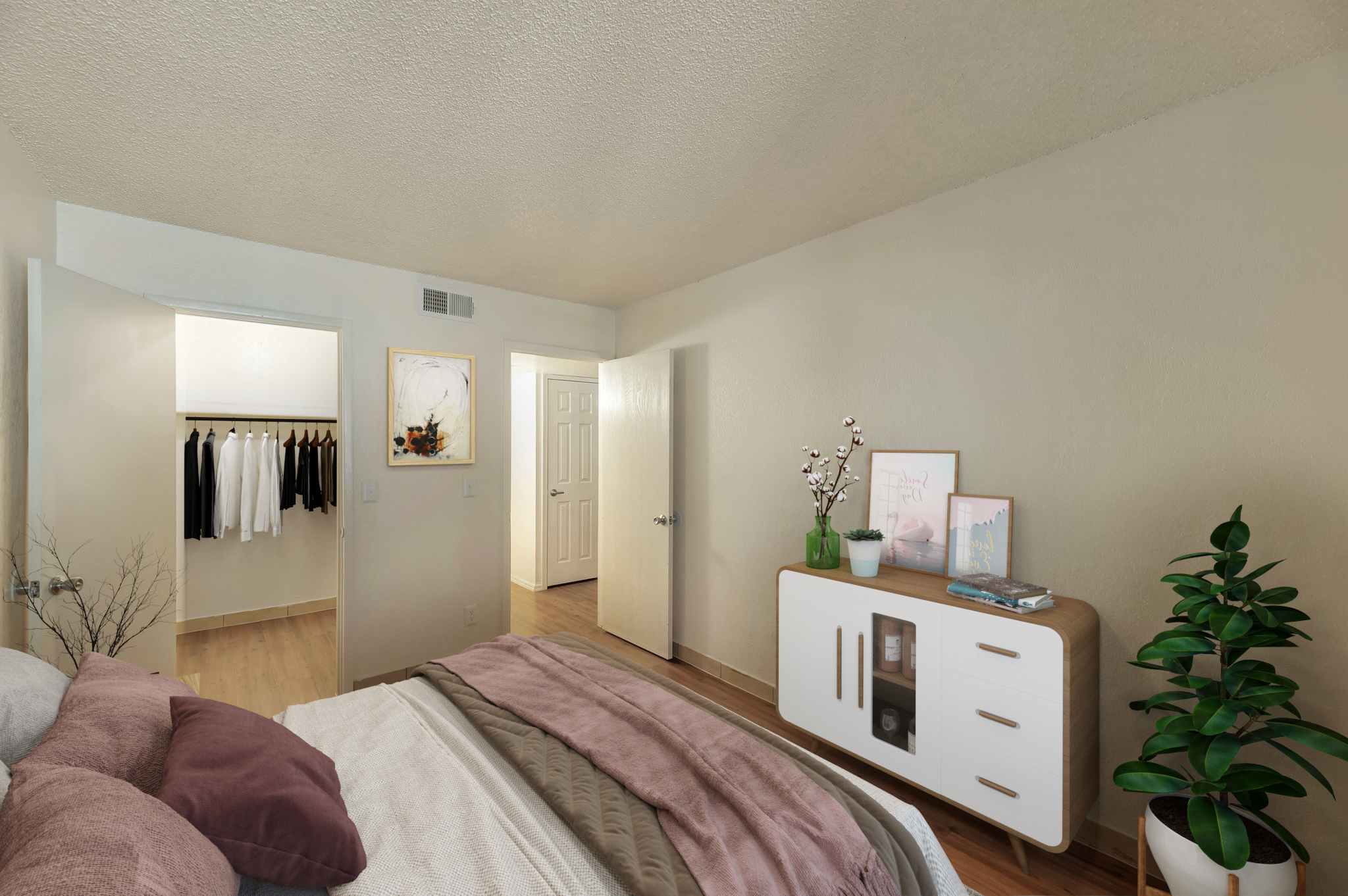 A cozy bedroom featuring a neatly made bed with decorative pillows, a nightstand with a plant, and a piece of wall art. A closet with hanging clothes is visible through an open door, alongside another door leading to a bathroom or additional space, all set against light-colored walls and wooden floors.