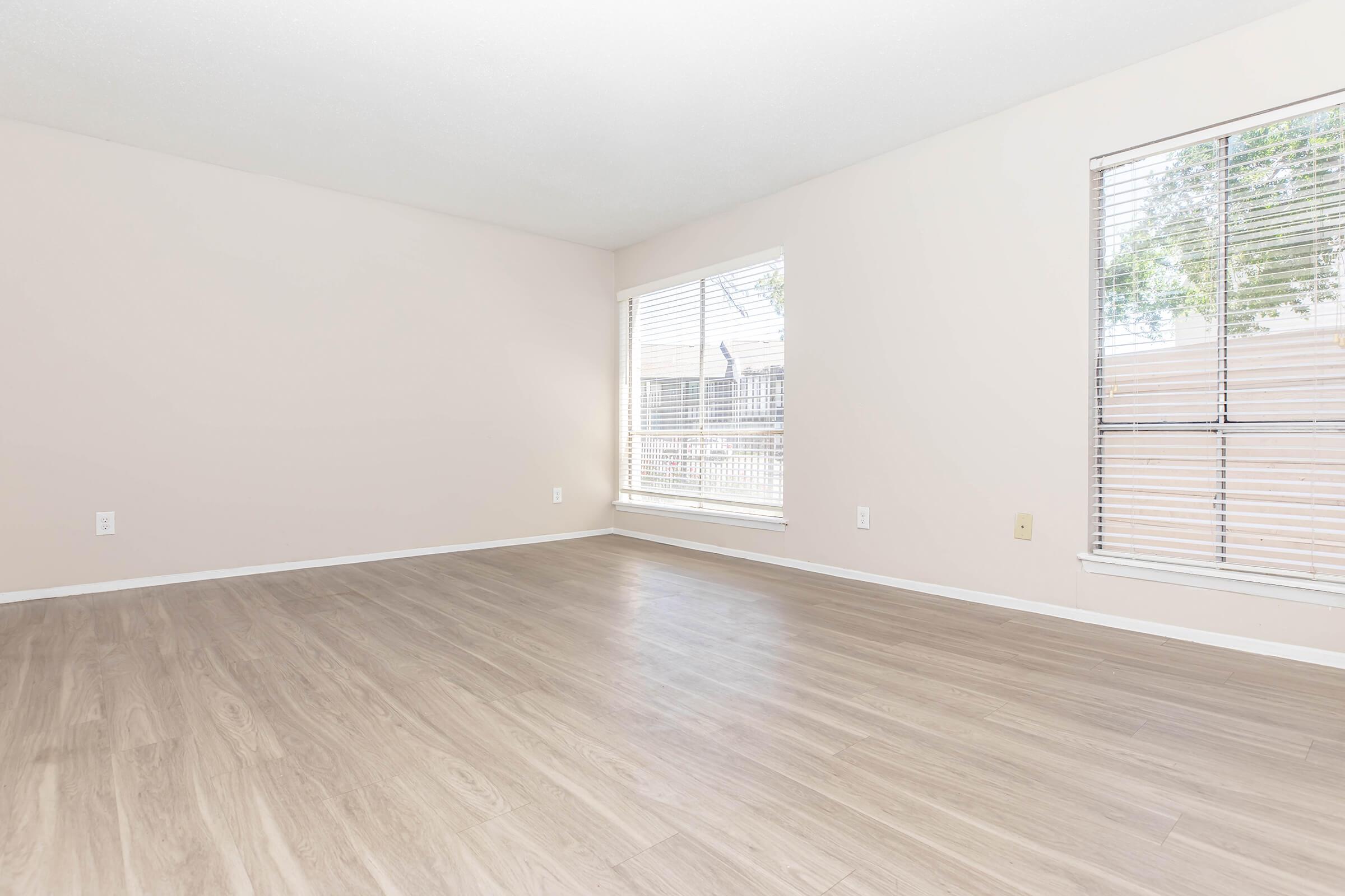 Spacious empty room with light-colored walls and wooden flooring. Natural light streams in through large windows with white blinds, creating a bright and inviting atmosphere. No furniture is present, emphasizing the openness of the space.