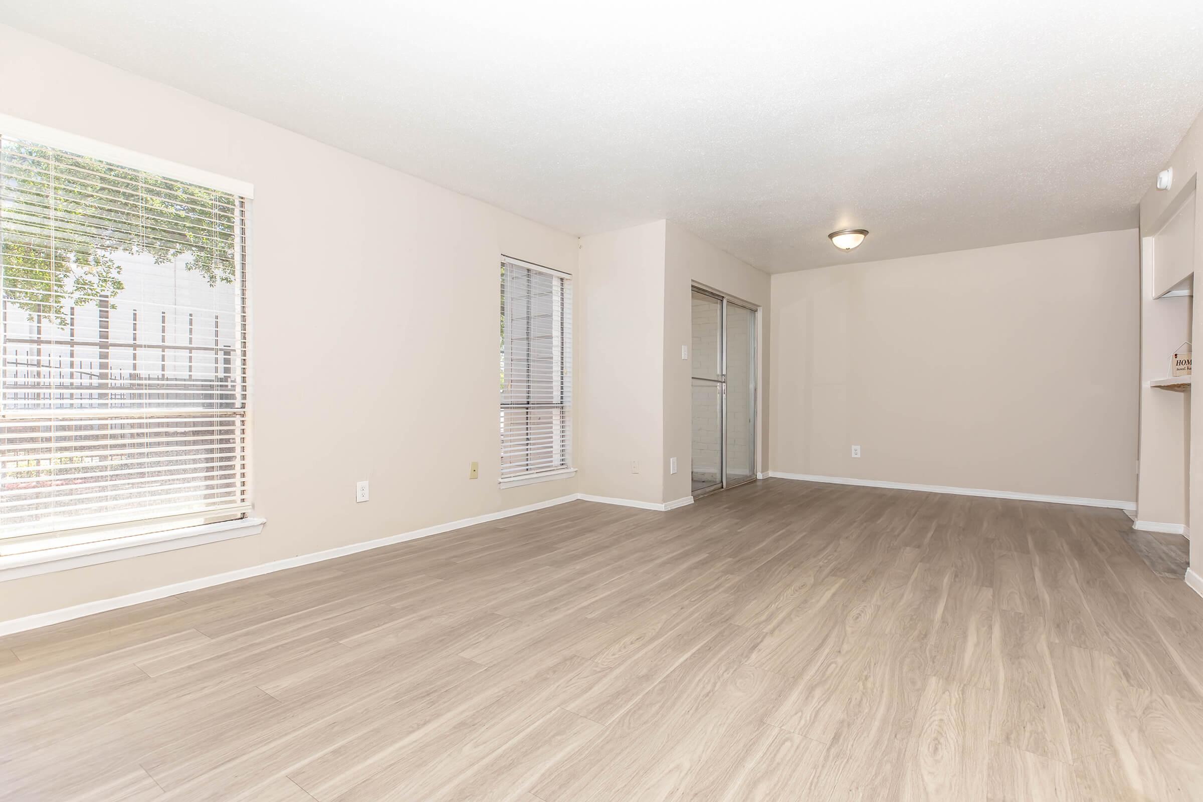 Spacious, empty living room featuring light-colored walls and wooden flooring. Large windows with blinds allow natural light to enter. The room has a ceiling light fixture and is adjacent to an area that appears to lead to a kitchen or dining space.