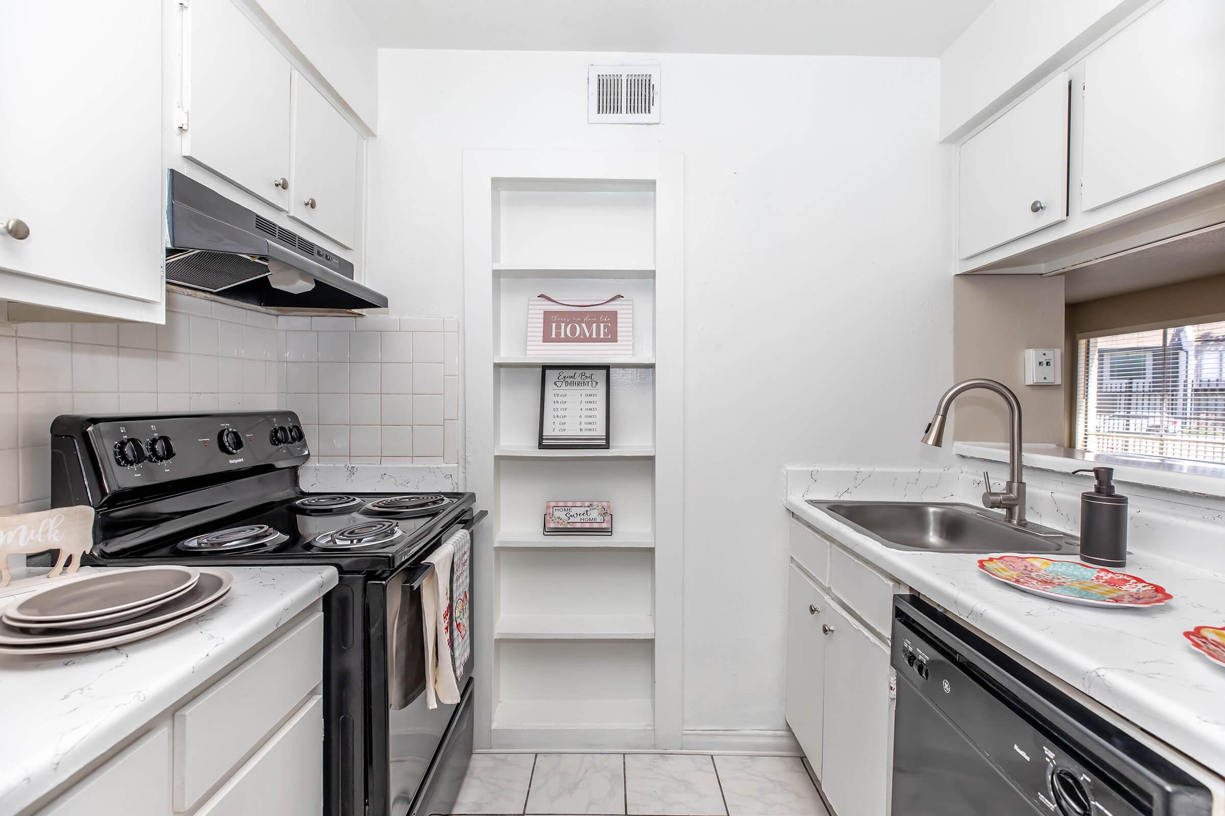 A modern kitchen featuring a black stove, white cabinetry, and a stainless steel sink. On the counter, there are decorative plates and a dark-colored soap dispenser. A shelf is built into the wall, displaying a decorative sign and home-related items. The floor is tiled in a light color.