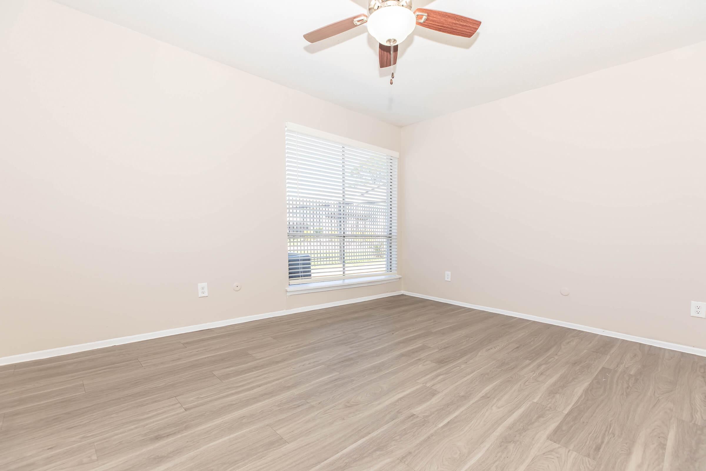 A spacious, empty room with light-colored walls and light wood flooring. A ceiling fan with wooden blades hangs in the center, and a large window with white blinds lets in natural light. The view outside shows grass, indicating a yard or outdoor area.