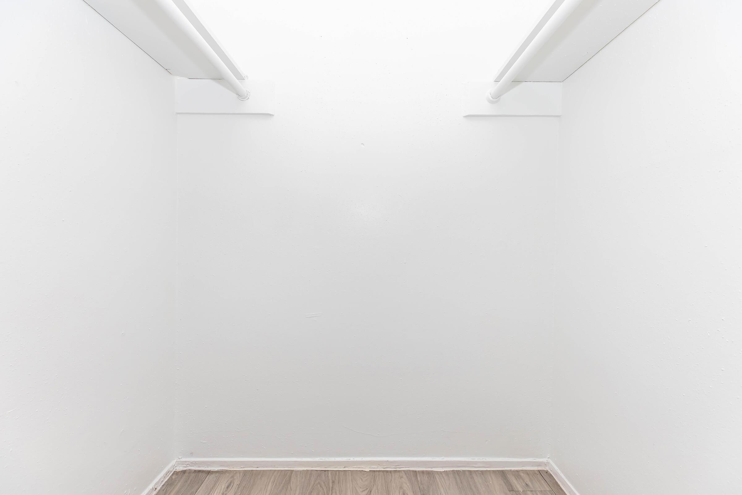 Empty white closet with two overhead rods and light wooden flooring, creating a minimalist and spacious appearance. The walls are smooth and unadorned, emphasizing simplicity and cleanliness.