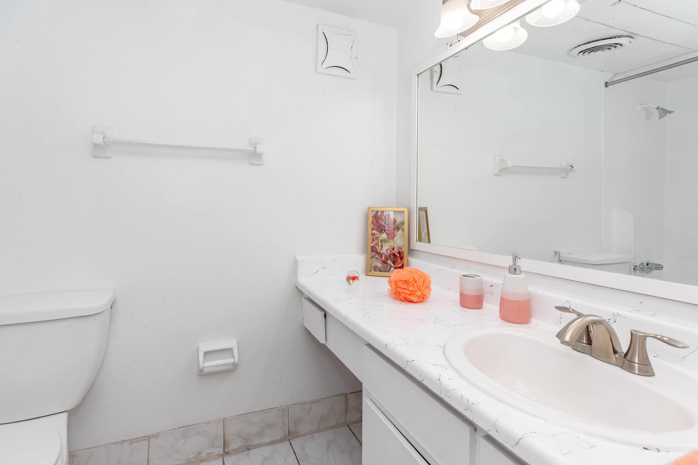 A clean and modern bathroom featuring a white countertop with a sink, a mirror above, and decorative items including a peach-colored accessory and a small plant. There is a toilet on the left and a towel rack on the wall, with light-colored tiles on the floor. Bright lighting enhances the spacious feel.