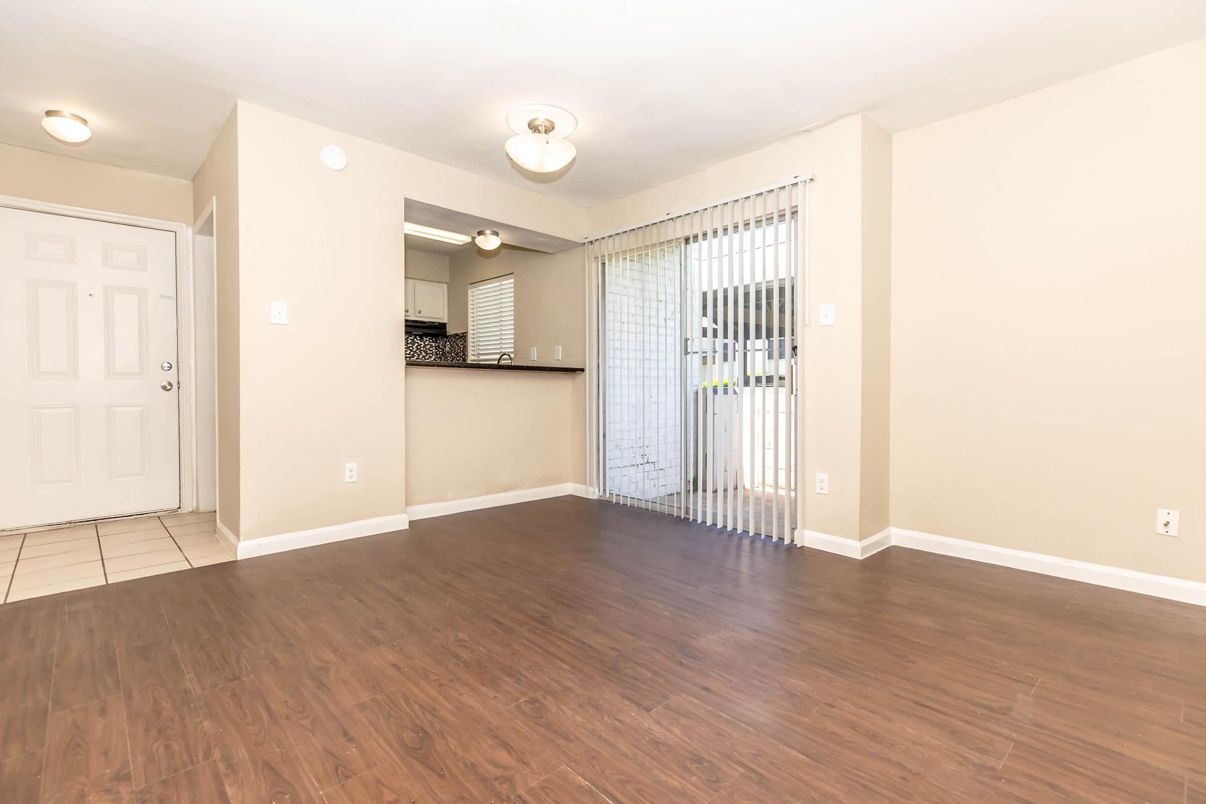 Spacious living area with light-colored walls and dark wood flooring. A door leads to an outdoor space, and there's a kitchen area visible through an open pass-through. Natural light brightens the room, which features a modern light fixture and sliding vertical blinds.