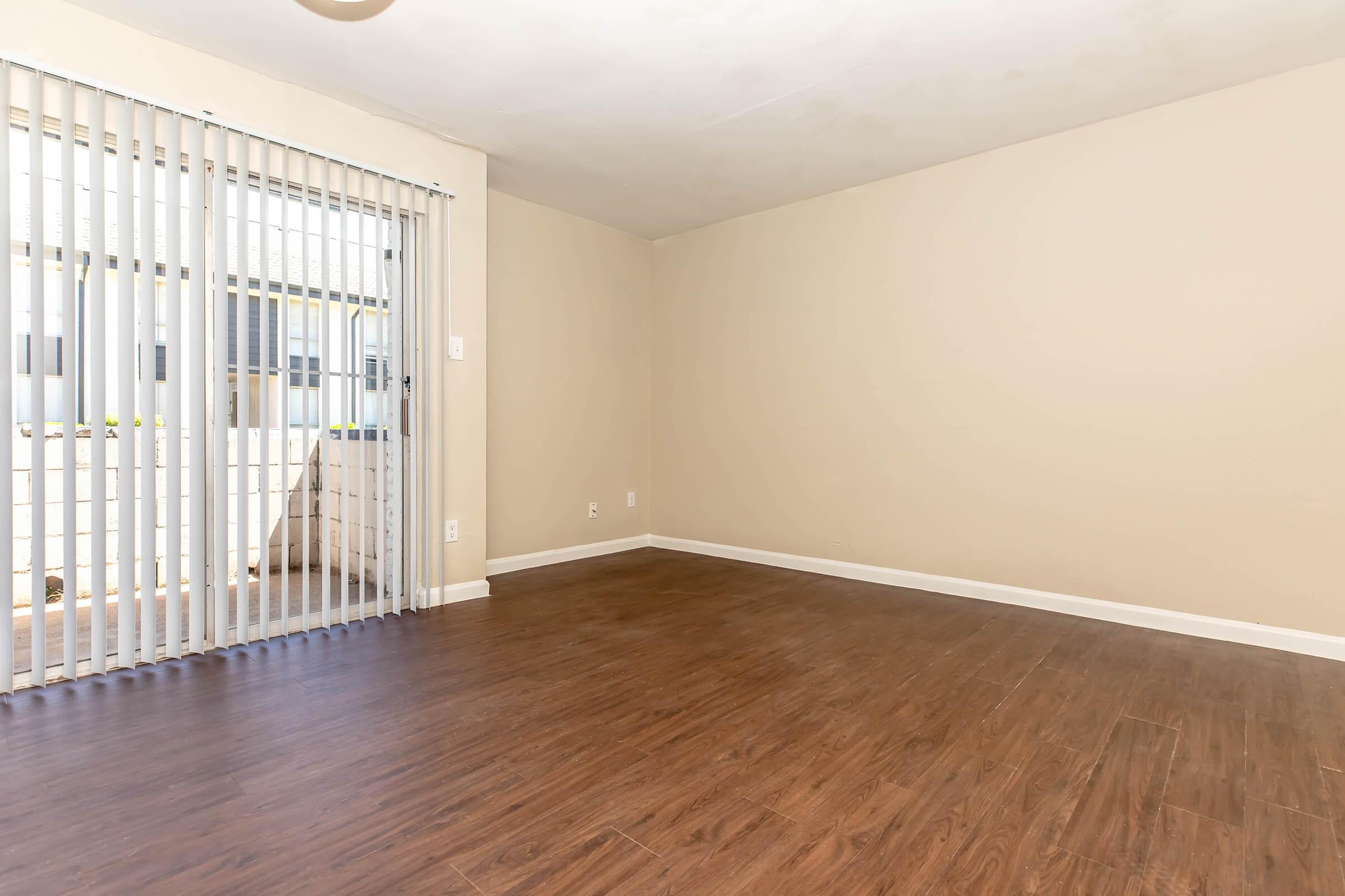 An empty room with light-colored walls and a wooden floor. To one side, there are vertical blinds partially covering a window. The space is bright and open, suggesting potential for decoration and furniture arrangement.