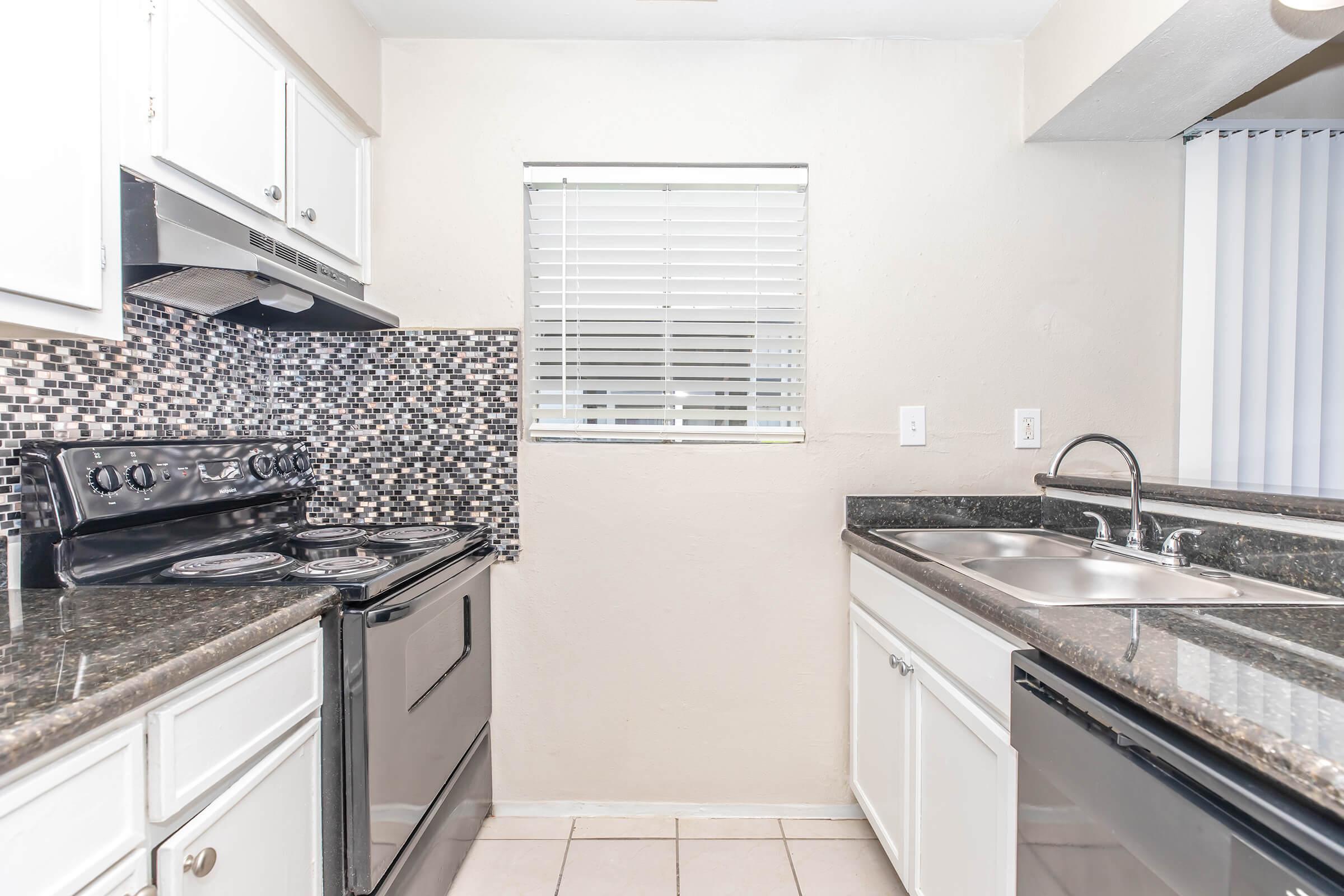 Modern kitchen featuring black and stainless steel appliances, including a gas stove and a double sink. The countertops are dark granite, and there's a decorative mosaic tile backsplash. Natural light enters through a window covered by blinds. The cabinets are white, creating a bright and clean atmosphere.