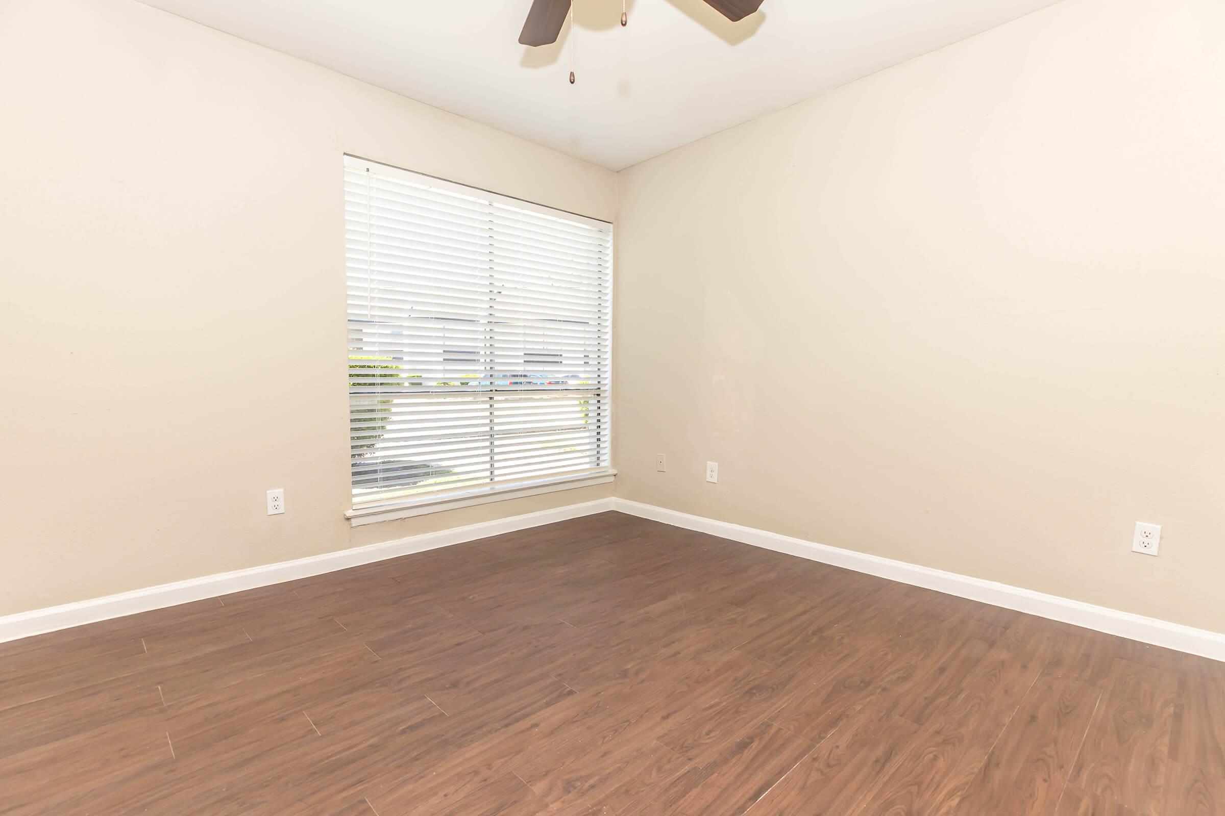 A light-colored room with hardwood flooring, featuring a ceiling fan and a large window with white blinds. The walls are painted a neutral shade, creating a bright and open feeling. The room is empty, allowing for various decoration and furniture arrangements.