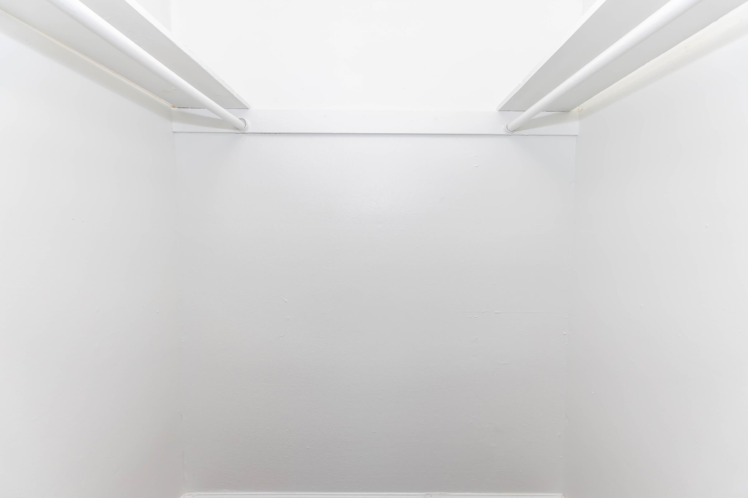 A clean, empty closet with white walls and two white hanging rods positioned near the top. The floor is bare, giving a minimalist and spacious appearance. Natural light brightens the interior, creating a fresh and inviting atmosphere.
