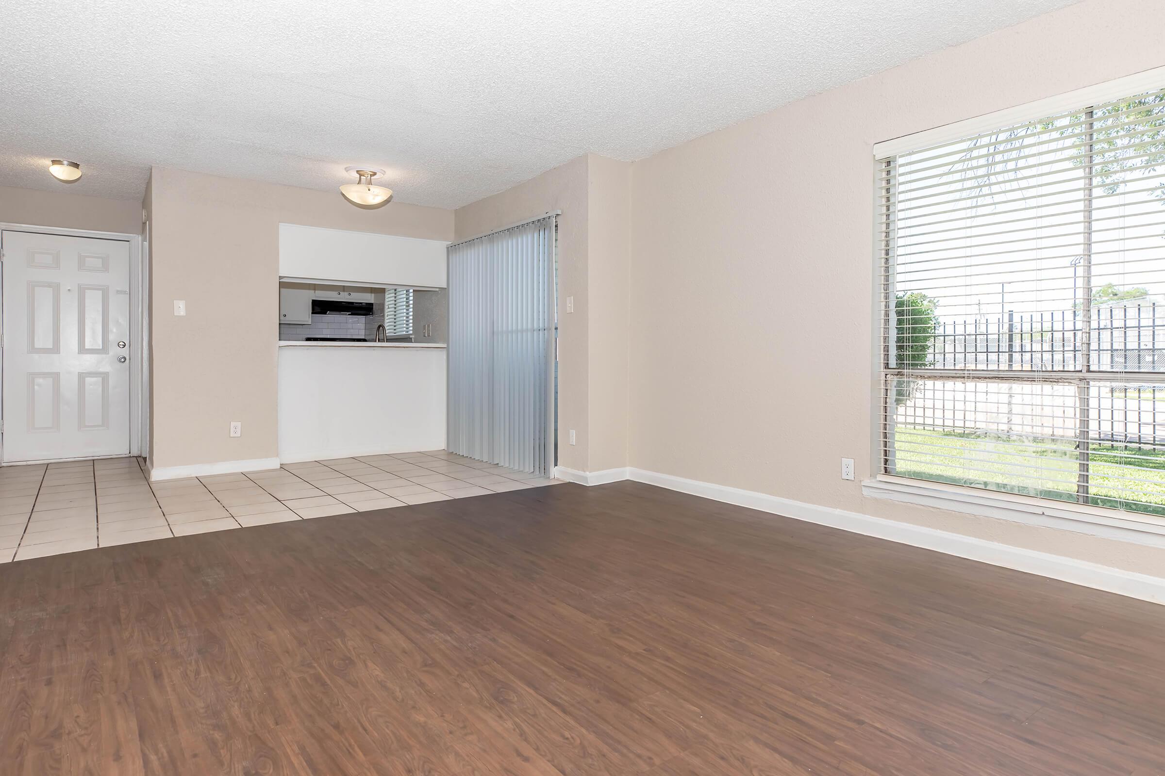 Spacious living area with light-colored walls and wood-style flooring, featuring a large window with blinds, providing a view of the outdoors. In the background, there is a kitchen area with a breakfast bar, and an entryway visible through a white door.