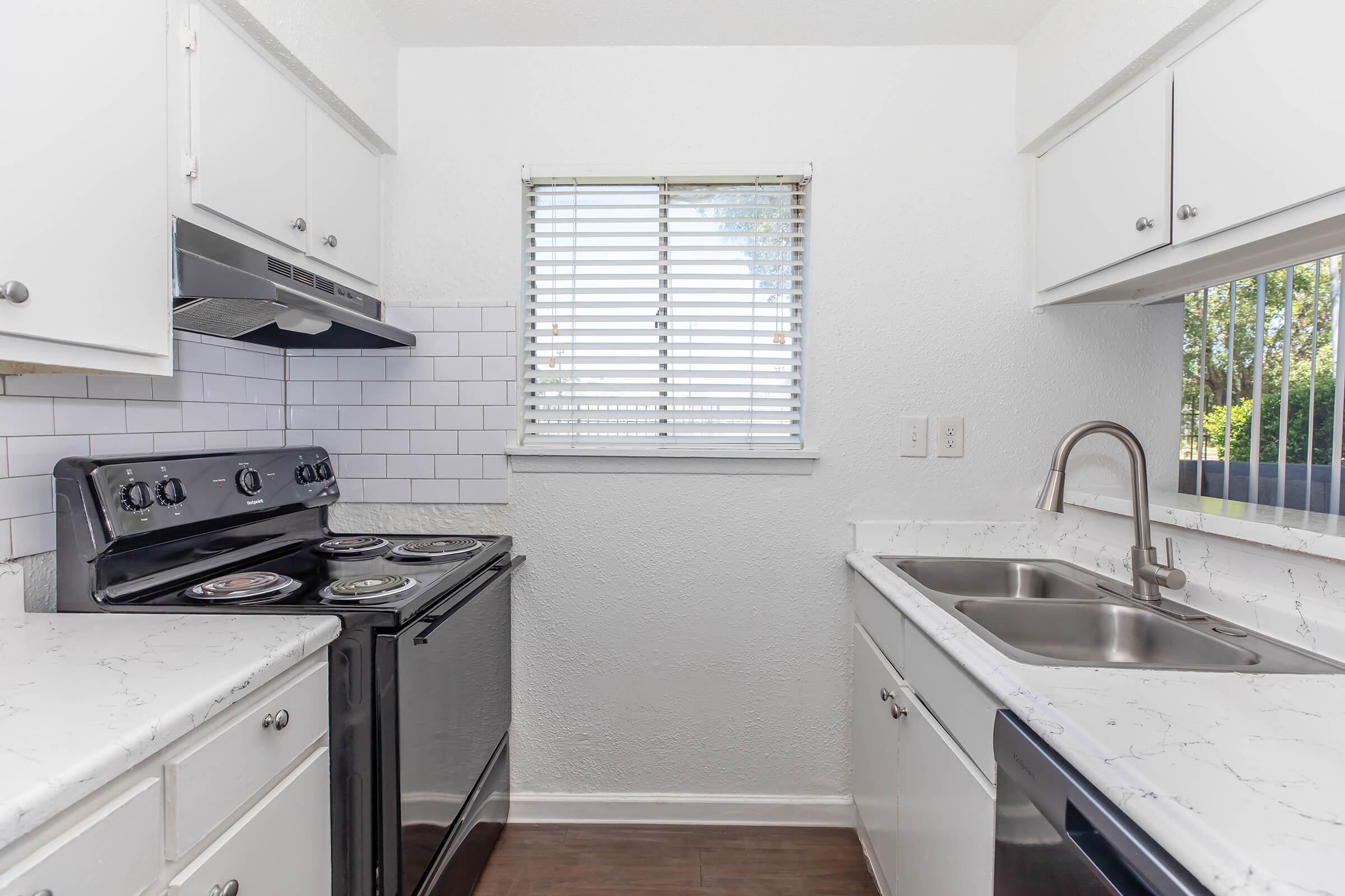 A modern kitchen featuring a white cabinetry, a black stove with an overhead vent, and a stainless steel sink. The countertops have a marbled design, and there is a window with blinds that provides natural light. The floor is a warm wood tone, creating a bright and inviting space.